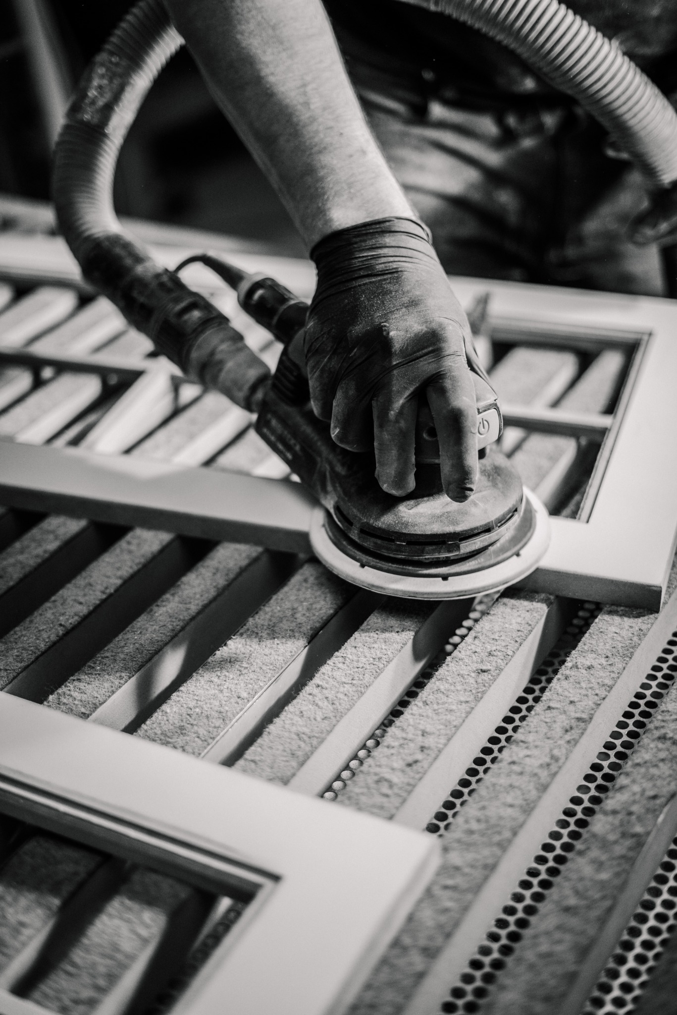Szlifowanie ramy okiennej szlifierką oscylacyjną z odsysaniem pyłu. Praca w warsztacie stolarskim, zbliżenie na dłoń w rękawicy i narzędzie. Monochromatyczne ujęcie.