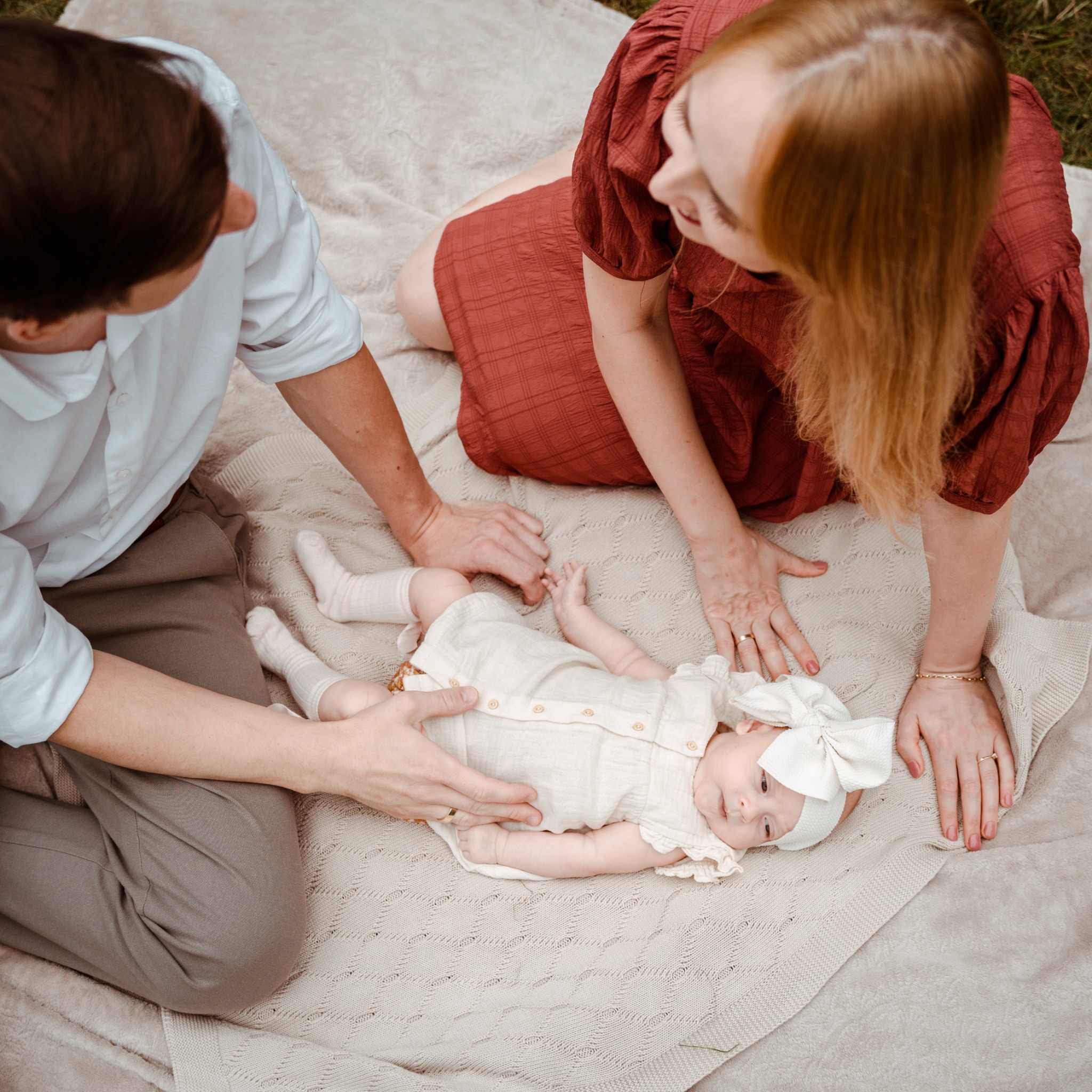 Rodzinne ujęcie z góry: rodzice czule spoglądają na leżącą na kocu córeczkę w kremowej sukience i z kokardą. Delikatne światło podkreśla bliskość i intymność chwili.