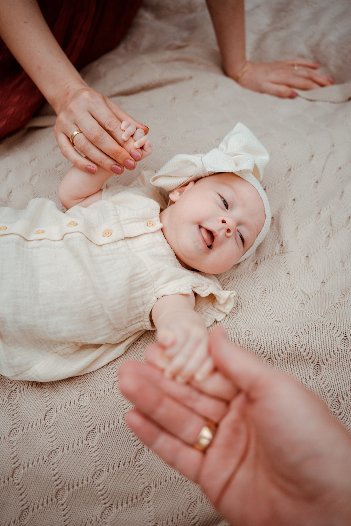 Urocze niemowlę w kremowym ubranku i opasce leży na beżowym kocyku, trzymając dłonie rodziców z widocznymi złotymi obrączkami. Ciepłe, naturalne światło.