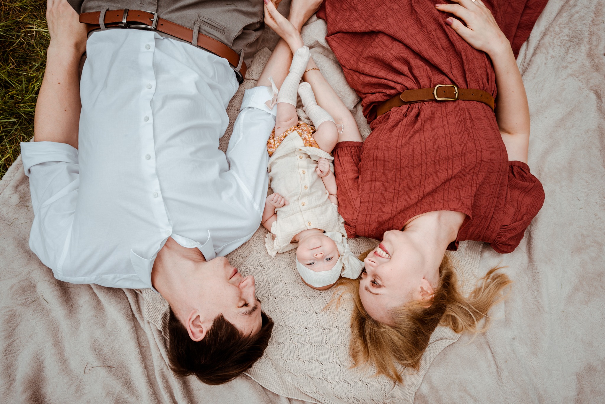 Rodzinna fotografia plenerowa: Mama w bordowej sukience, tata w bieli, a między nimi niemowlę w beżowym ubranku, leżący na kocu w trawie, patrzący w obiektyw.