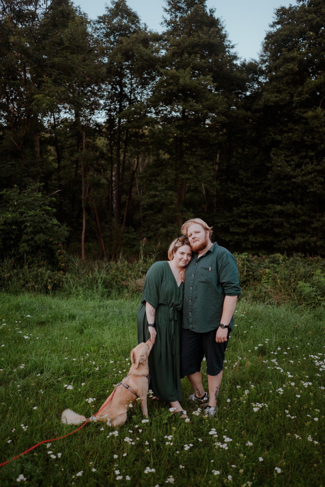Zakochana para w zielonych strojach pozuje z psem na łące pełnej białych kwiatów, w tle ciemny las. Romantyczna atmosfera, naturalne światło.