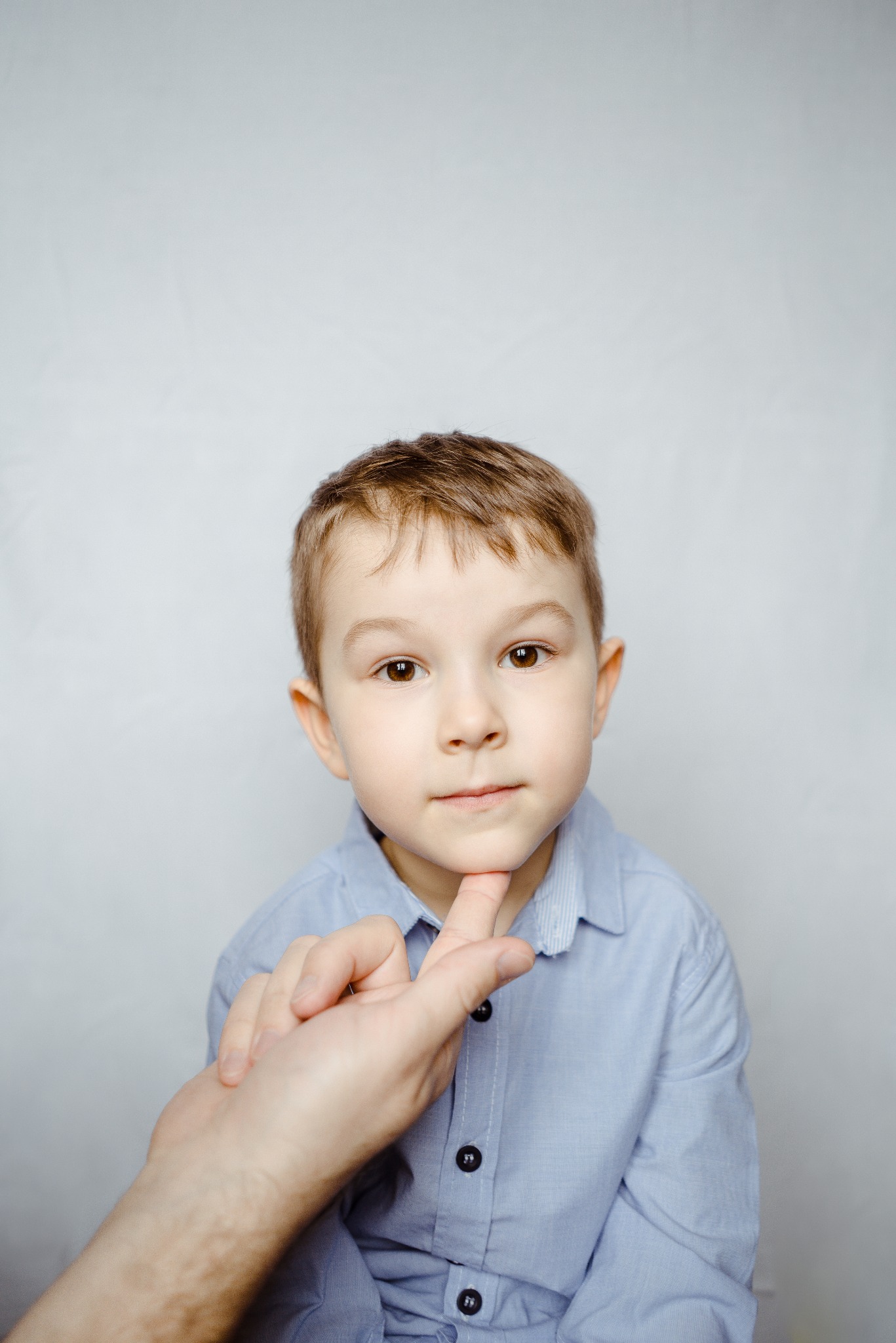 Portret chłopca w błękitnej koszuli, z dłonią podtrzymującą jego podbródek, na jasnym tle. Ujęcie z bliska, skupione na twarzy dziecka.