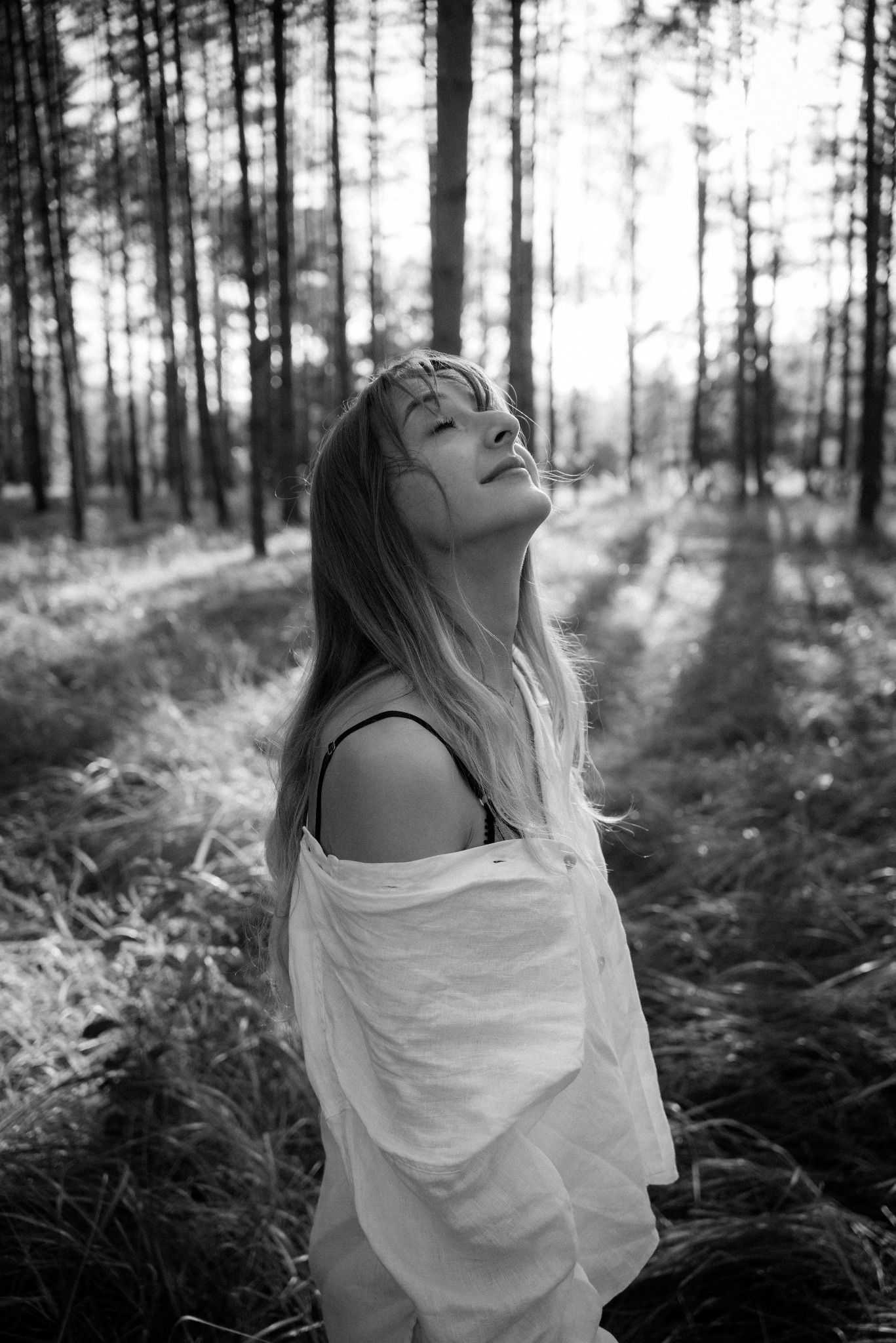 Czarno-biała fotografia kobiety z uniesioną głową, w rozpiętej koszuli na tle lasu, z promieniami słońca przebijającymi przez drzewa. Artystyczne ujęcie.