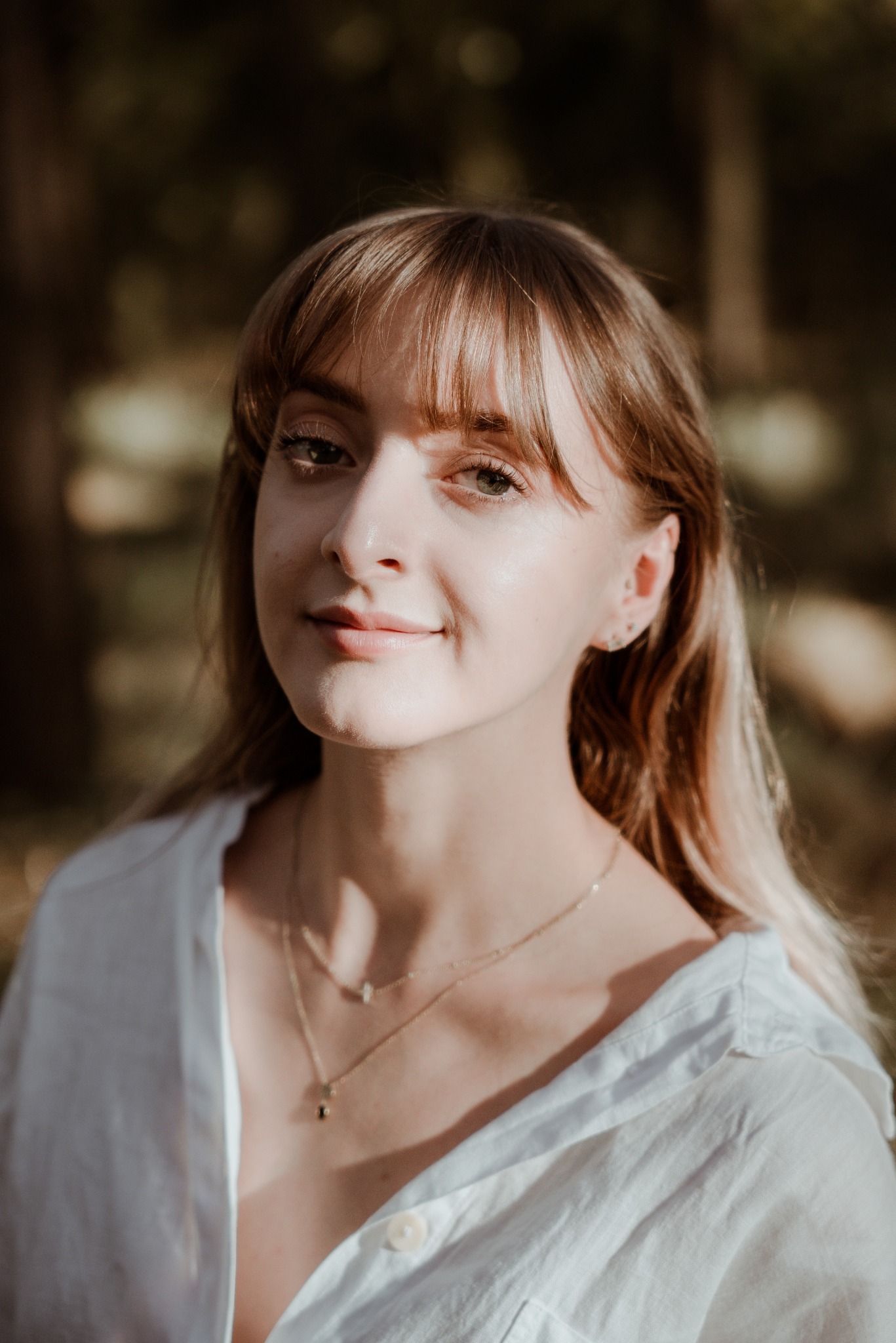 Portret młodej kobiety z blond włosami i grzywką, ubranej w białą koszulę, z delikatnym uśmiechem, na tle rozmytej zieleni. Naturalne światło podkreśla rysy twarzy.