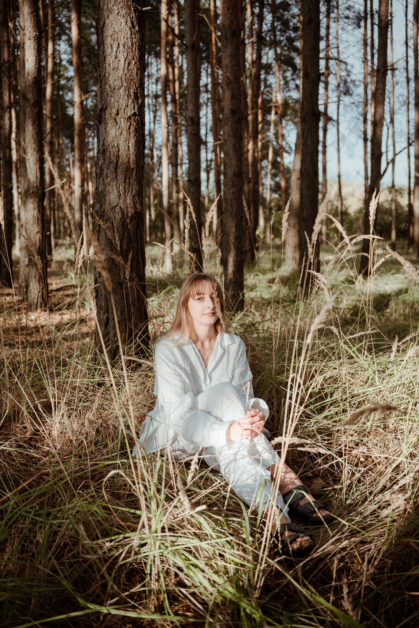 Kobieta w bieli siedzi w trawie na tle lasu sosnowego. Naturalne światło, delikatny uśmiech, swobodna poza. Kadrowanie od kolan w górę, z bliska.