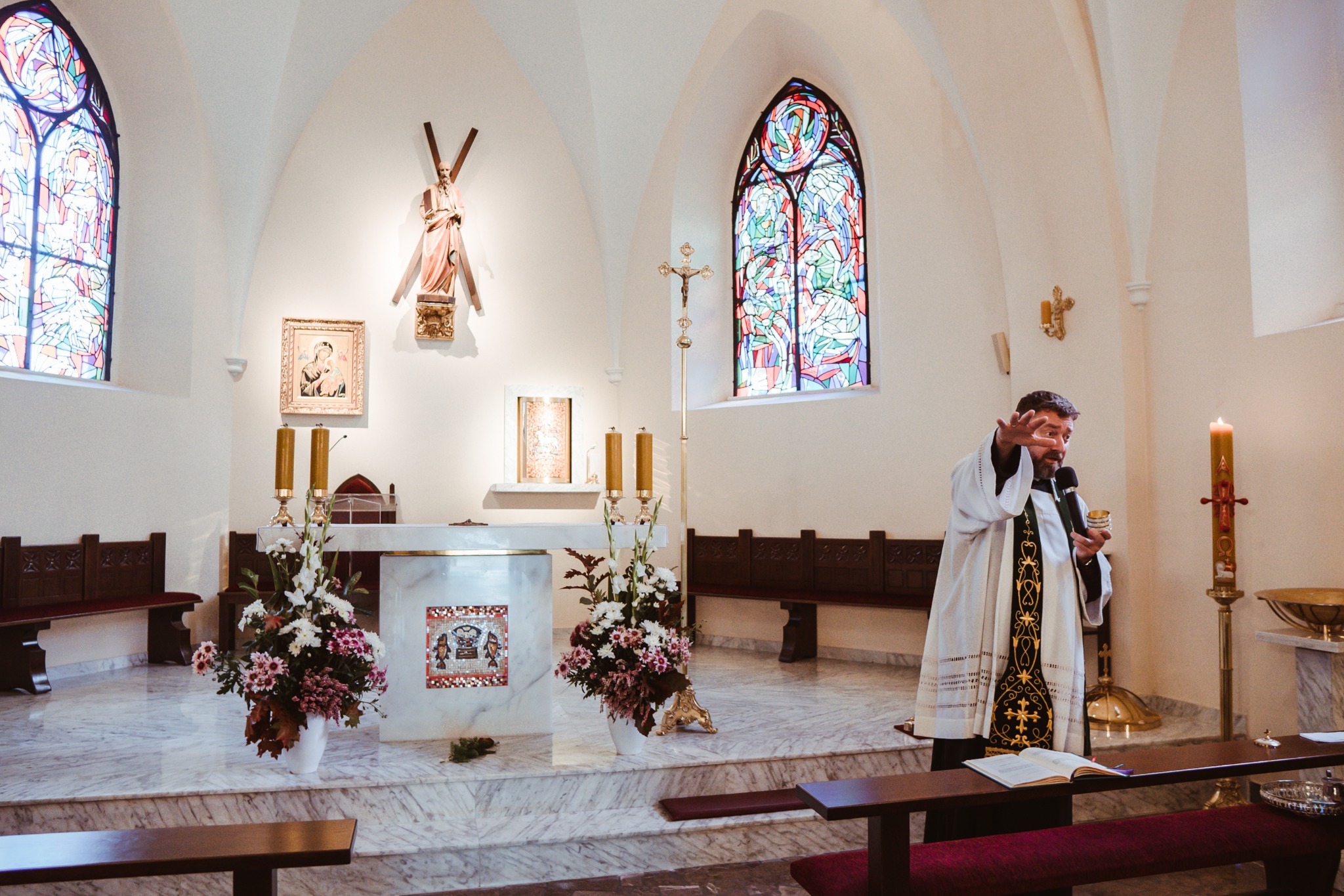 Ksiądz z mikrofonem w dłoni podczas ceremonii w kościele. W tle ołtarz z kwiatami, witraże i ławki. Ujęcie z boku, wnętrze kościoła oświetlone naturalnym światłem.