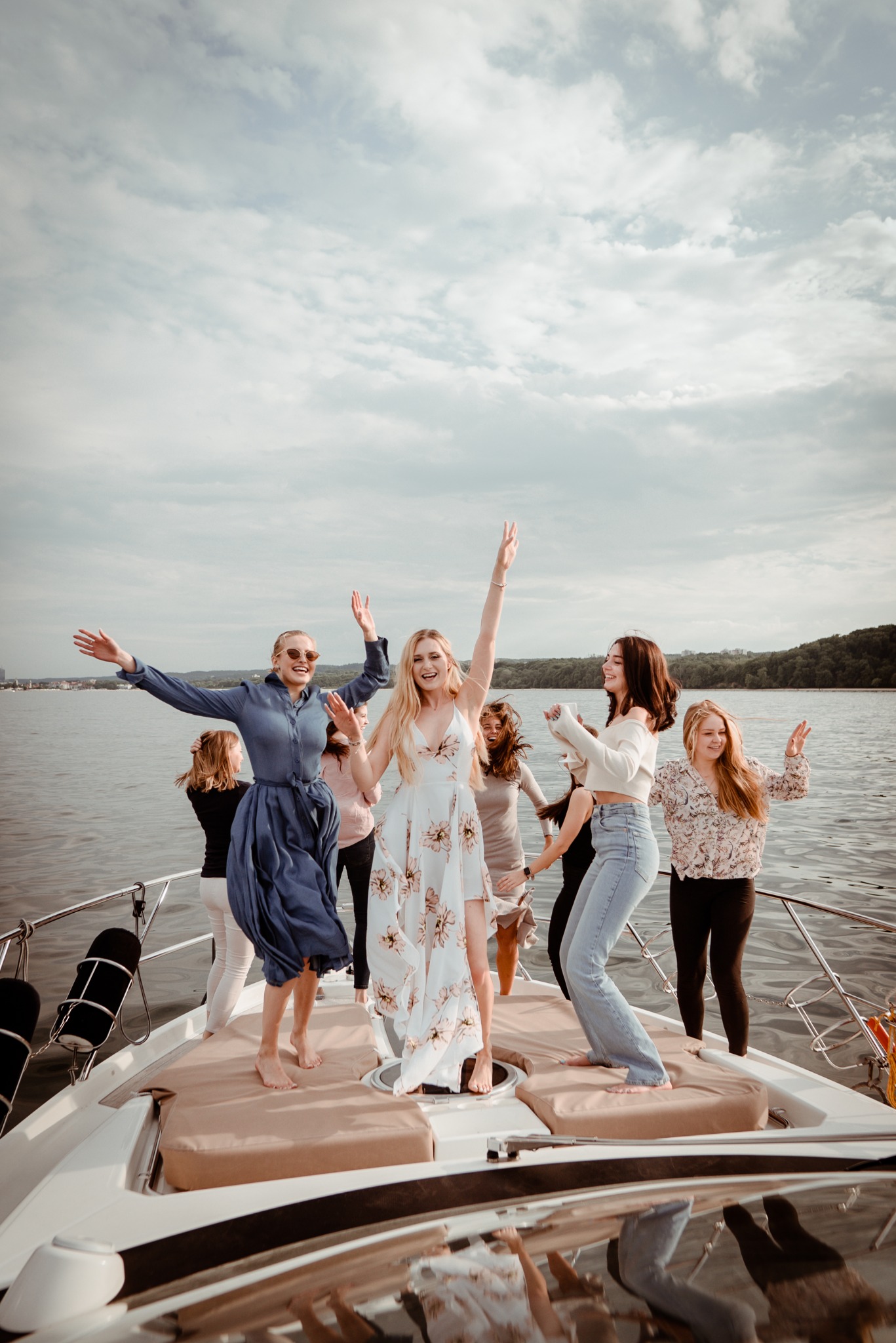 Grupa kobiet świętuje na jachcie, w tle woda i pochmurne niebo. Uśmiechnięte, rozbawione osoby w ruchu, w letnich stylizacjach. Fotografia z perspektywy dziobu łodzi.