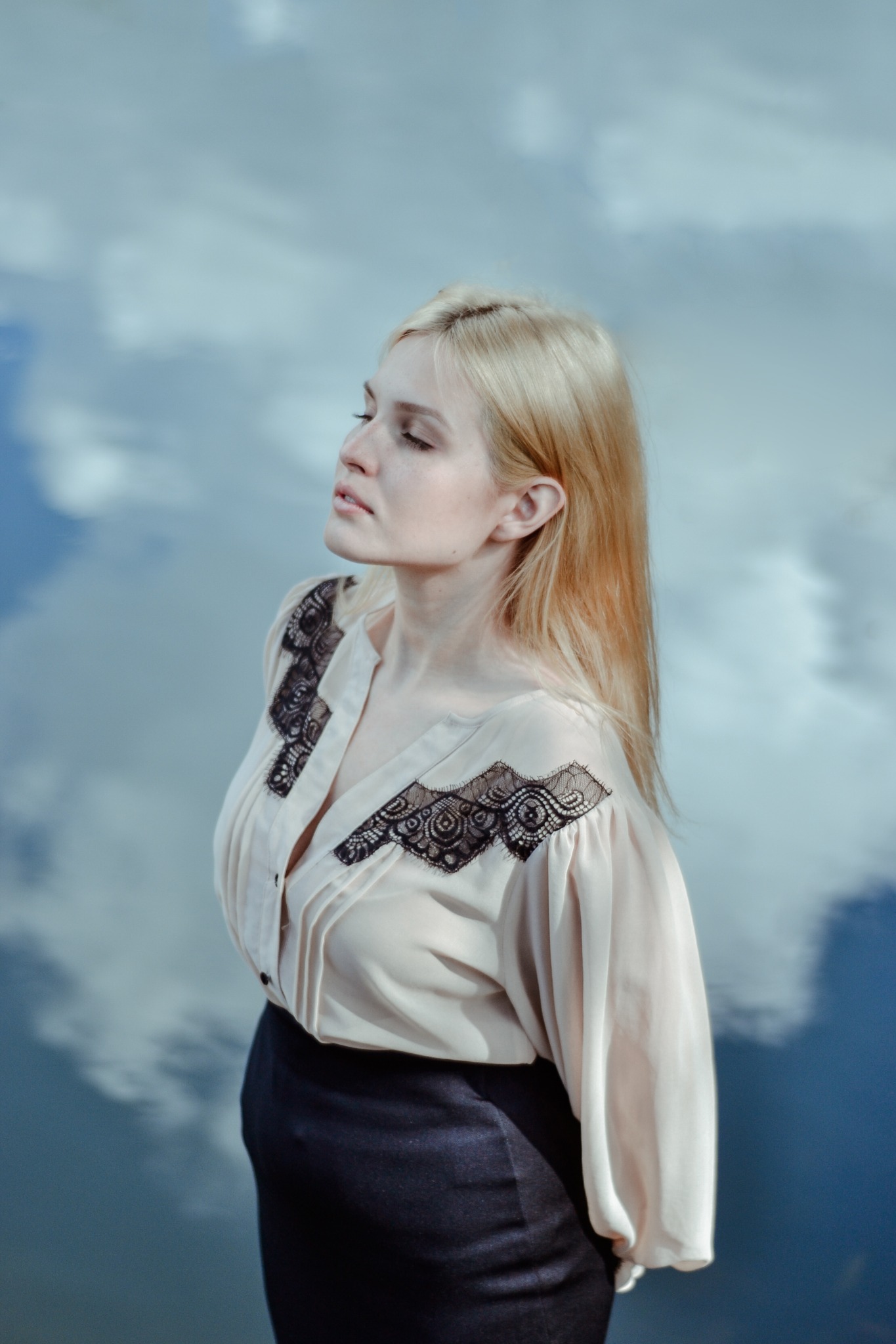 Portret kobiety o jasnych włosach, w bluzce z koronkowymi wstawkami, na tle nieba odbijającego się w wodzie. Ujęcie z profilu, delikatne światło.