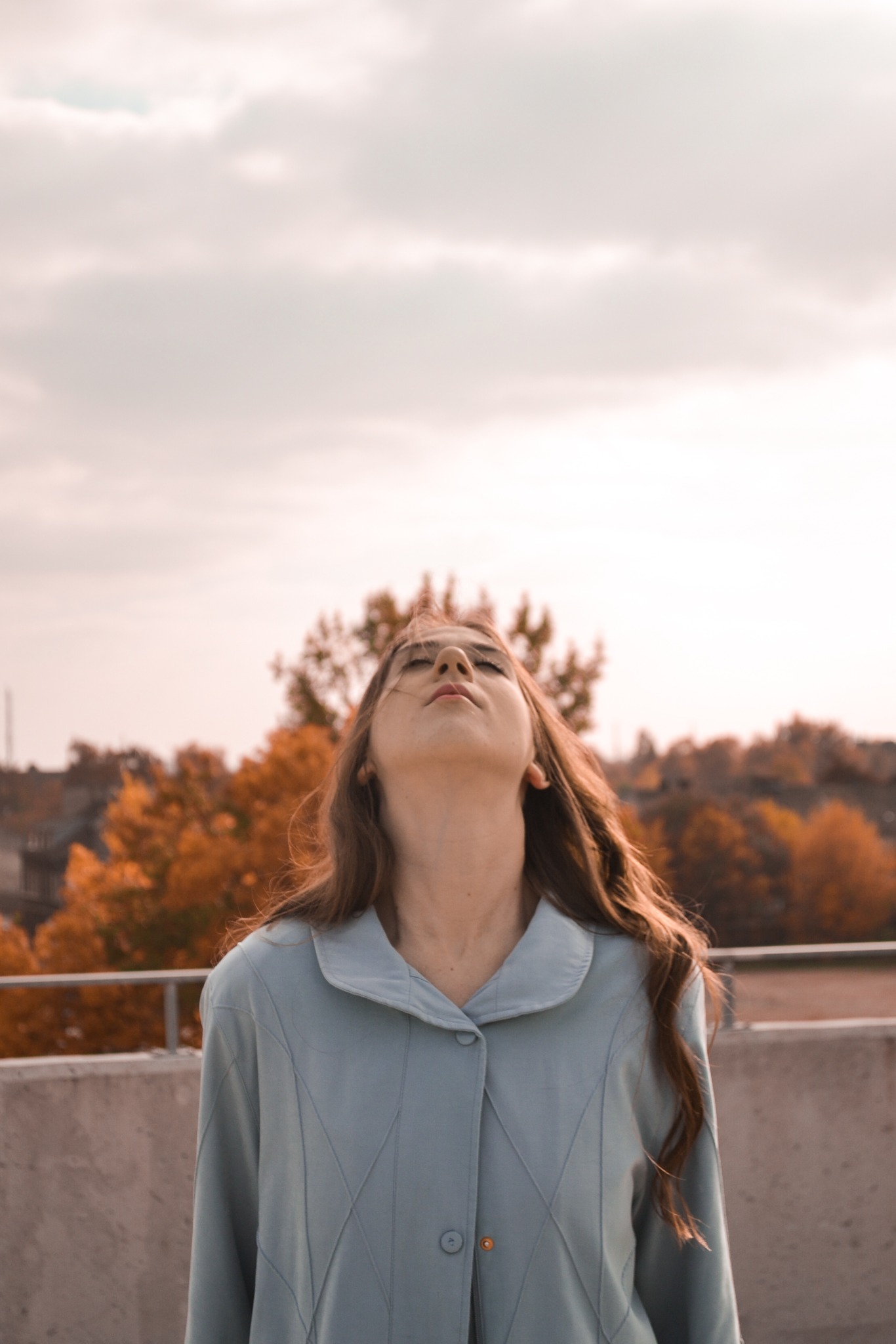 Portret kobiety z długimi włosami, w błękitnej kurtce, spoglądającej w górę na tle jesiennego krajobrazu i pochmurnego nieba. Ujęcie z dołu.