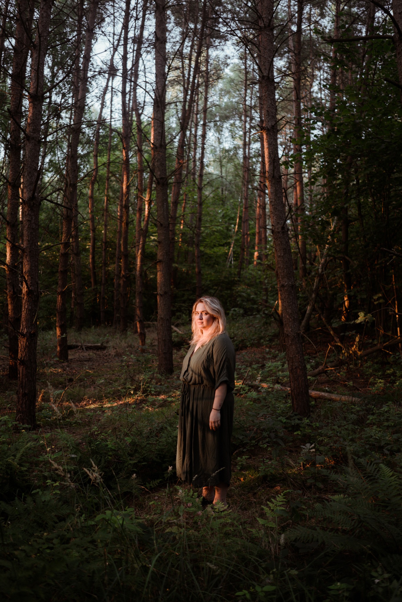 Portret kobiety w leśnej scenerii. Blond włosy, długa sukienka w odcieniach zieleni. Drzewa tworzą naturalną ramę, a delikatne światło podkreśla atmosferę tajemniczości.