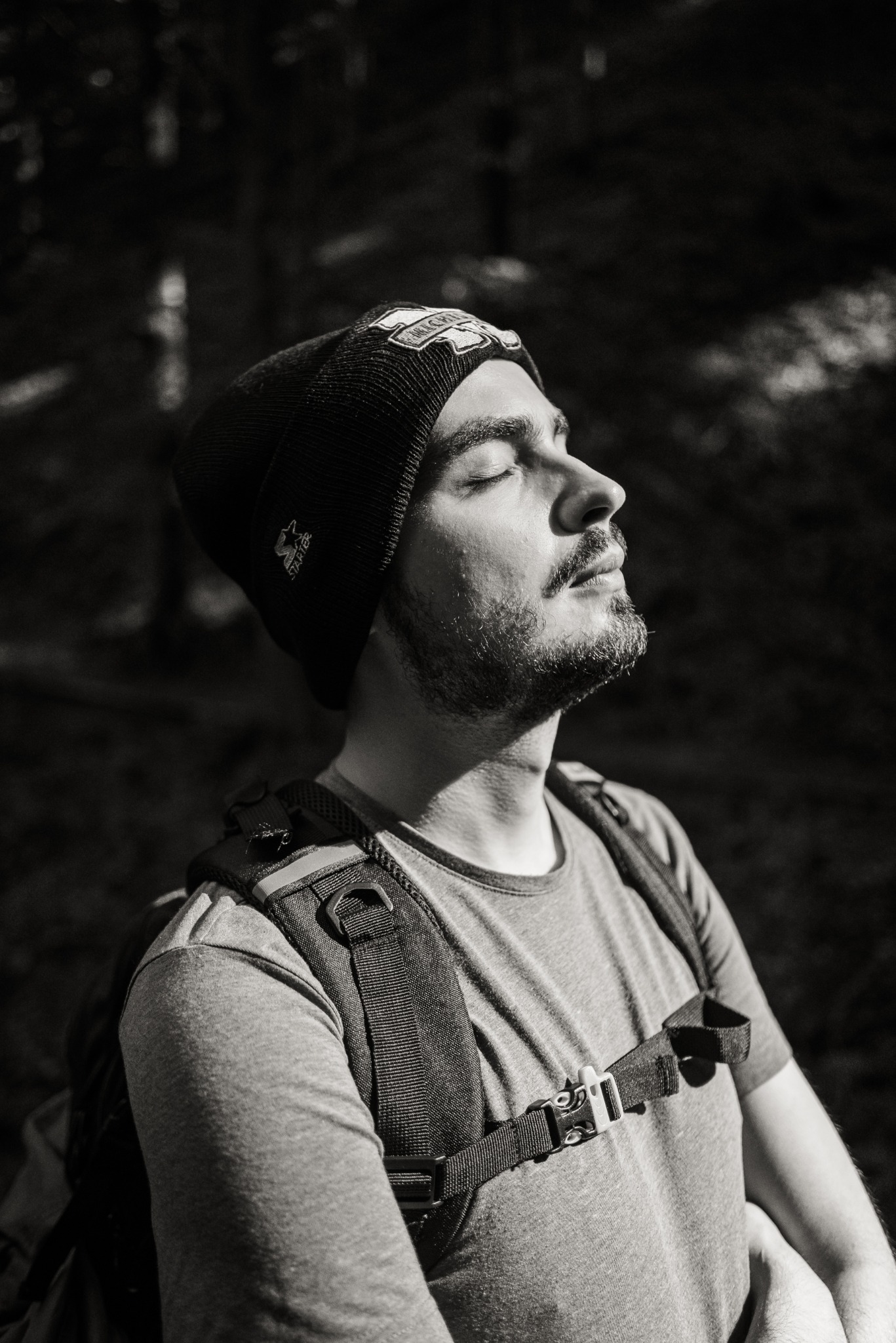 Czarno-białe zdjęcie mężczyzny z brodą w czapce i plecaku, z zamkniętymi oczami, skierowanego twarzą do słońca. Ujęcie z dołu, w tle ciemne drzewa.