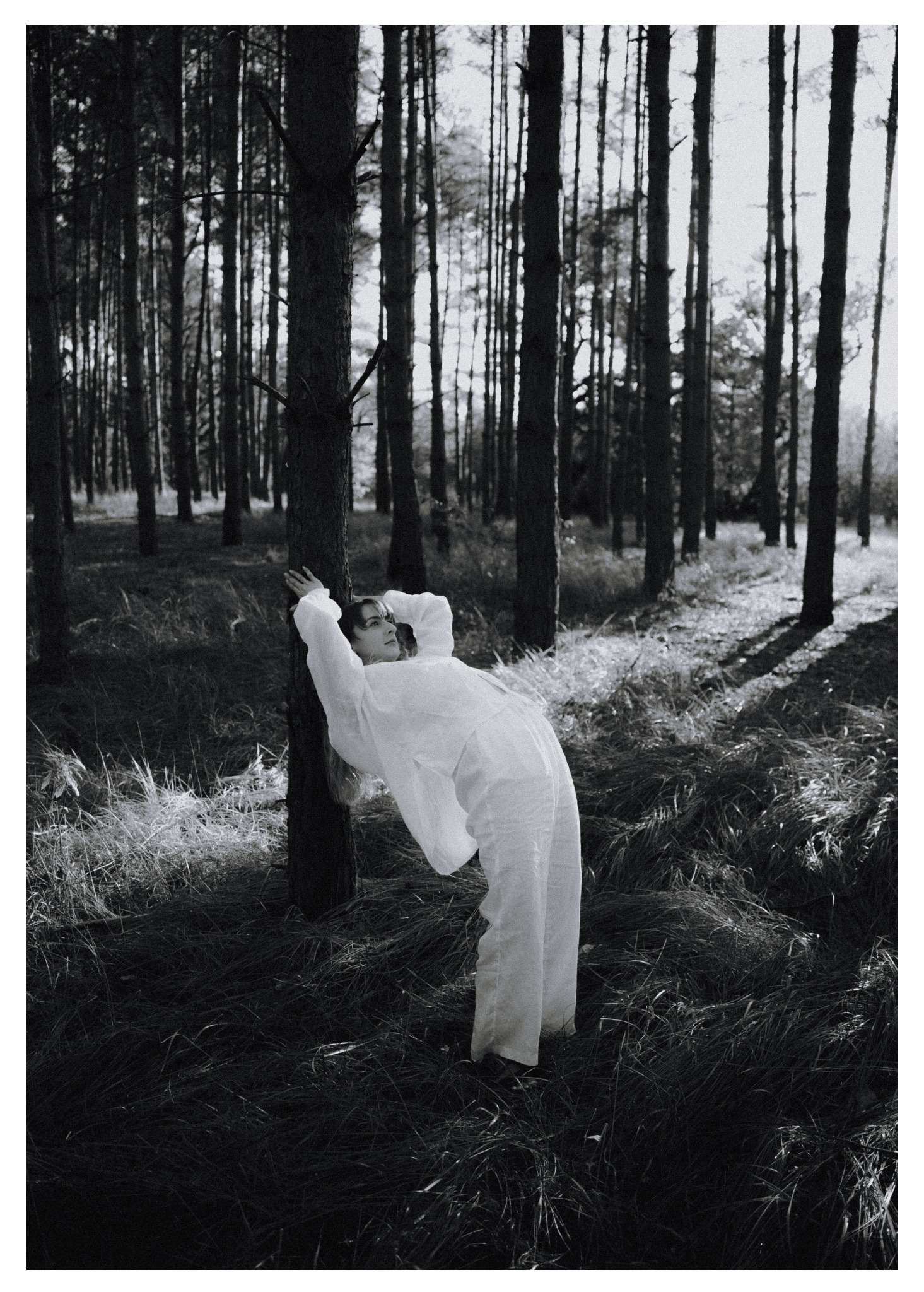 Czarno-biała sesja kobieca w lesie: kobieta w białym kombinezonie opiera się o drzewo, z zamyślonym wyrazem twarzy, otoczona wysokimi drzewami i trawą.