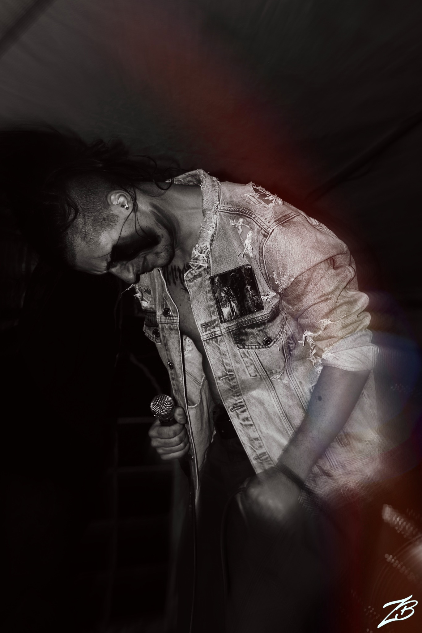 Czarno-białe zdjęcie wokalisty z mikrofonem podczas występu na scenie, w dżinsowej kurtce z naszywką, pochylonego w dynamicznym ruchu.