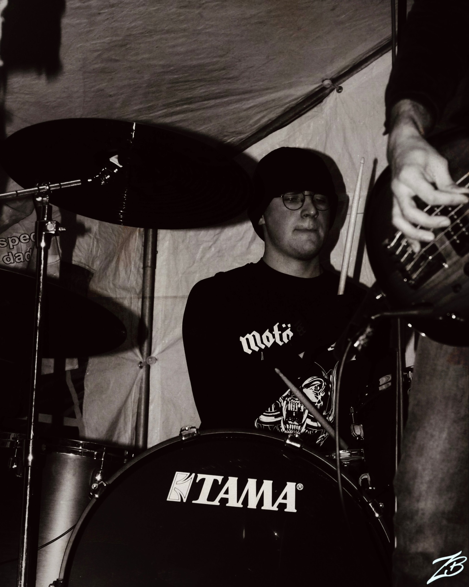 Czarno-białe zdjęcie perkusisty z Poznania podczas występu na scenie. Widoczny bębny marki TAMA i fragment gitary basowej. Ujęcie z bliska, dynamiczne oświetlenie.