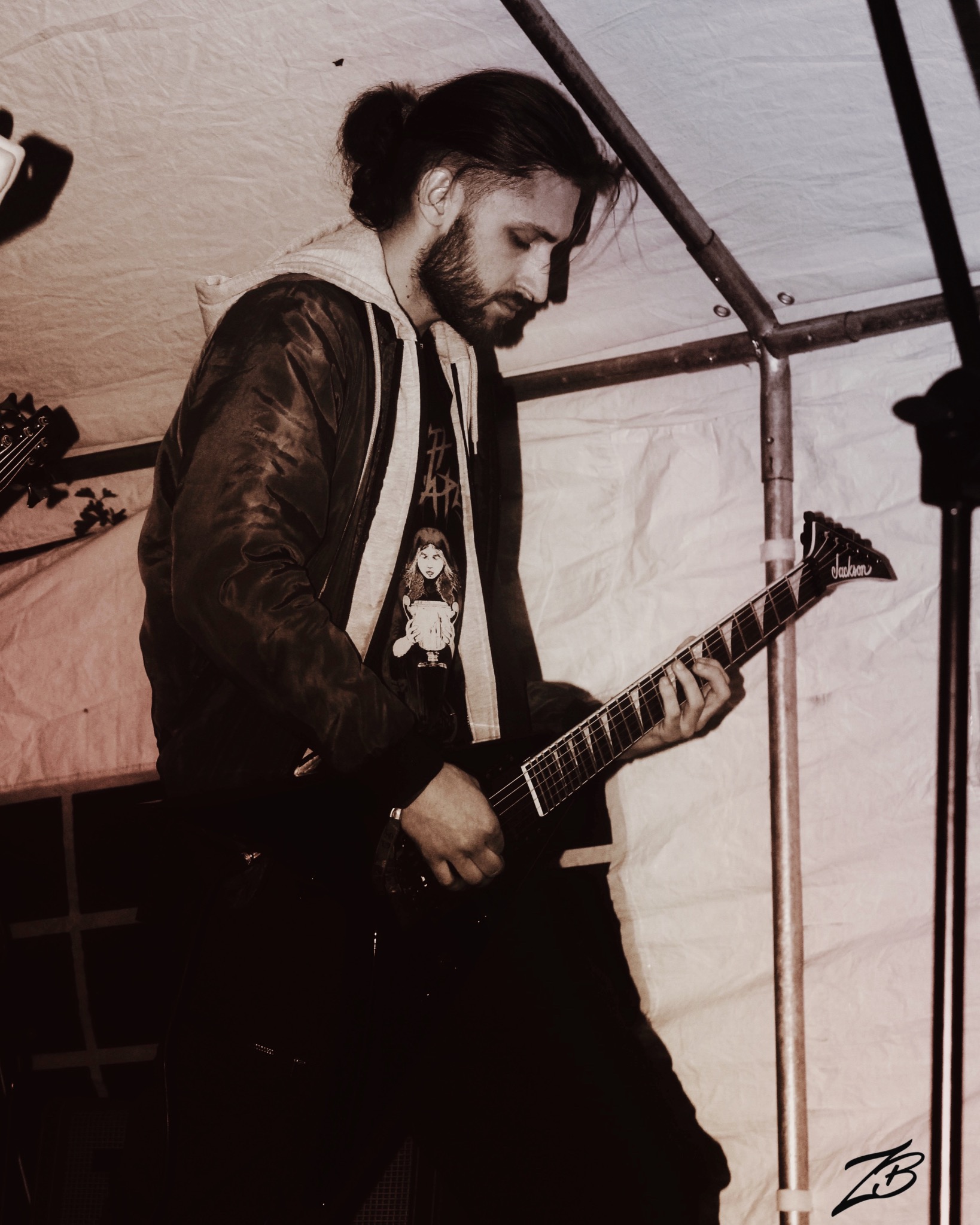 Muzyk z brodą i włosami spiętymi w kok, grający na czarnej gitarze elektrycznej Jackson podczas koncertu pod namiotem. Ubrany w kurtkę i koszulkę z nadrukiem.