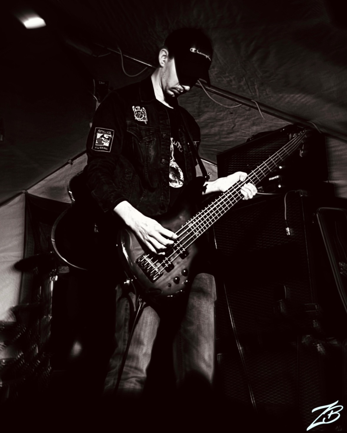 Czarno-białe zdjęcie basisty grającego na basie podczas koncertu, z widocznymi wzmacniaczami i elementami perkusji w tle. Uwagę przykuwa naszywka Metallica na kurtce muzyka.