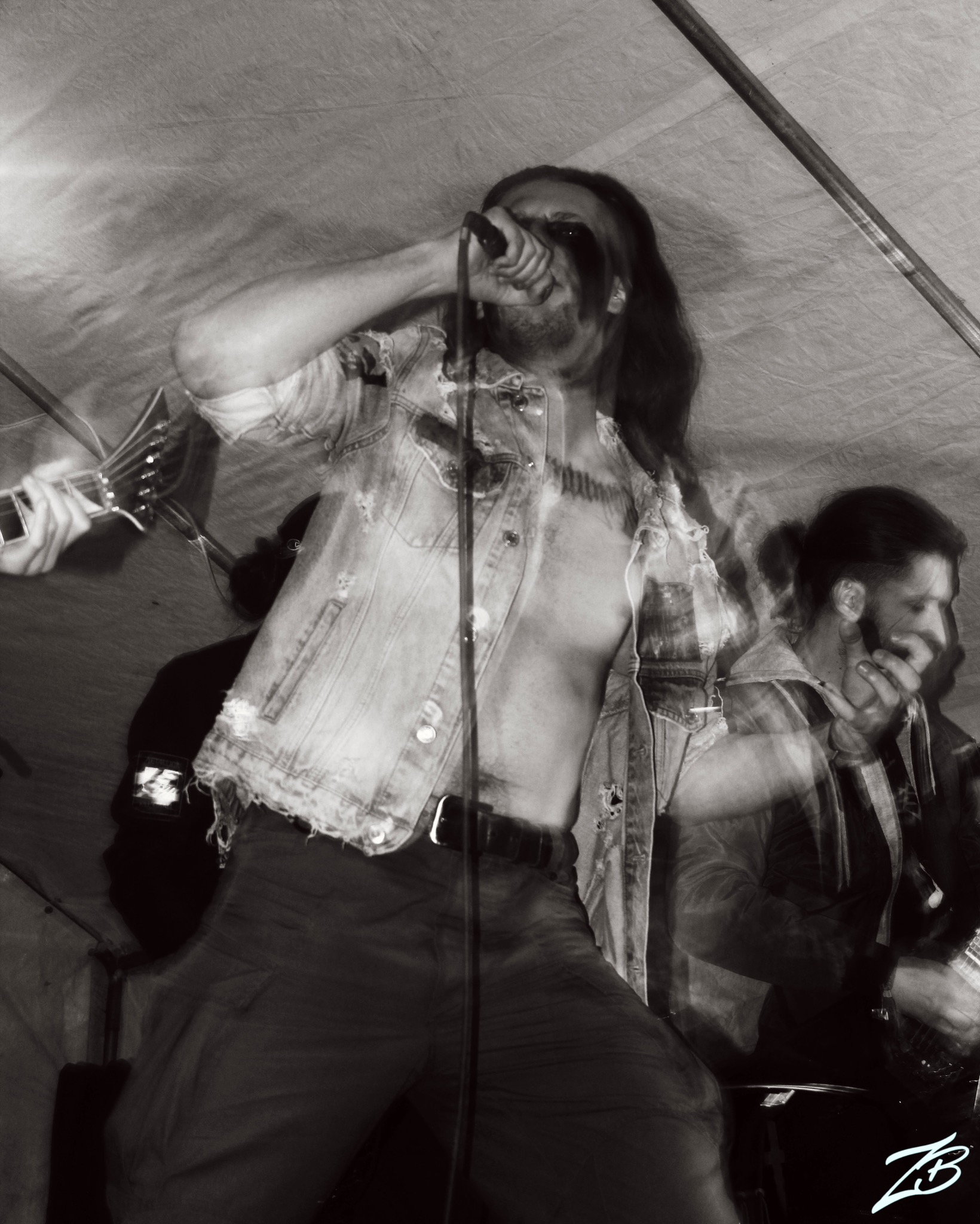 Czarno-białe zdjęcie wokalisty z długimi włosami i makijażem scenicznym, śpiewającego do mikrofonu podczas koncertu. W tle rozmyte sylwetki muzyków i instrumentów.