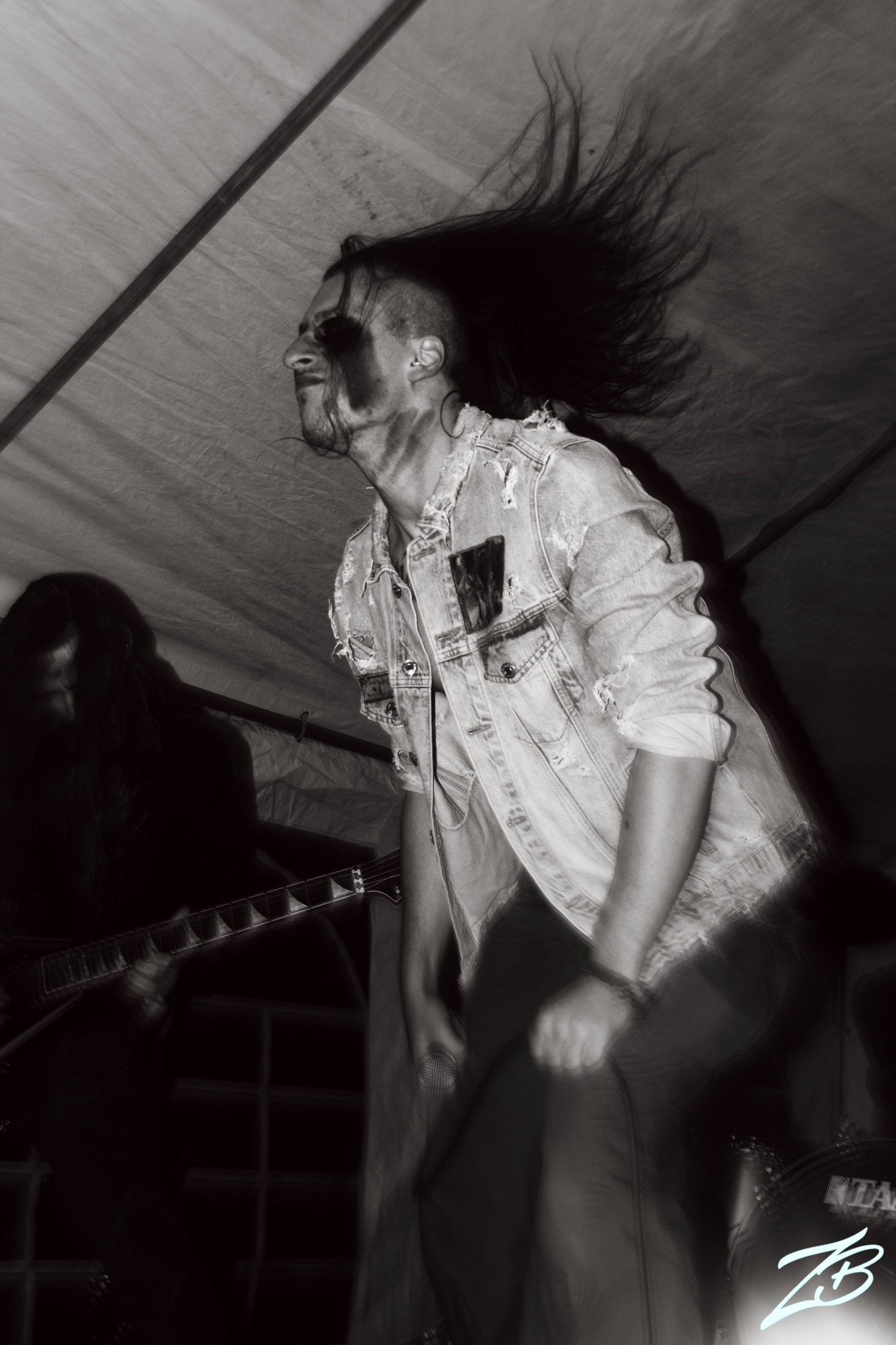 Czarno-białe zdjęcie wokalisty z długimi włosami i gitarzysty podczas koncertu rockowego. Wokalista w dżinsowej kurtce, ekspresyjny ruch, dynamiczne ujęcie.