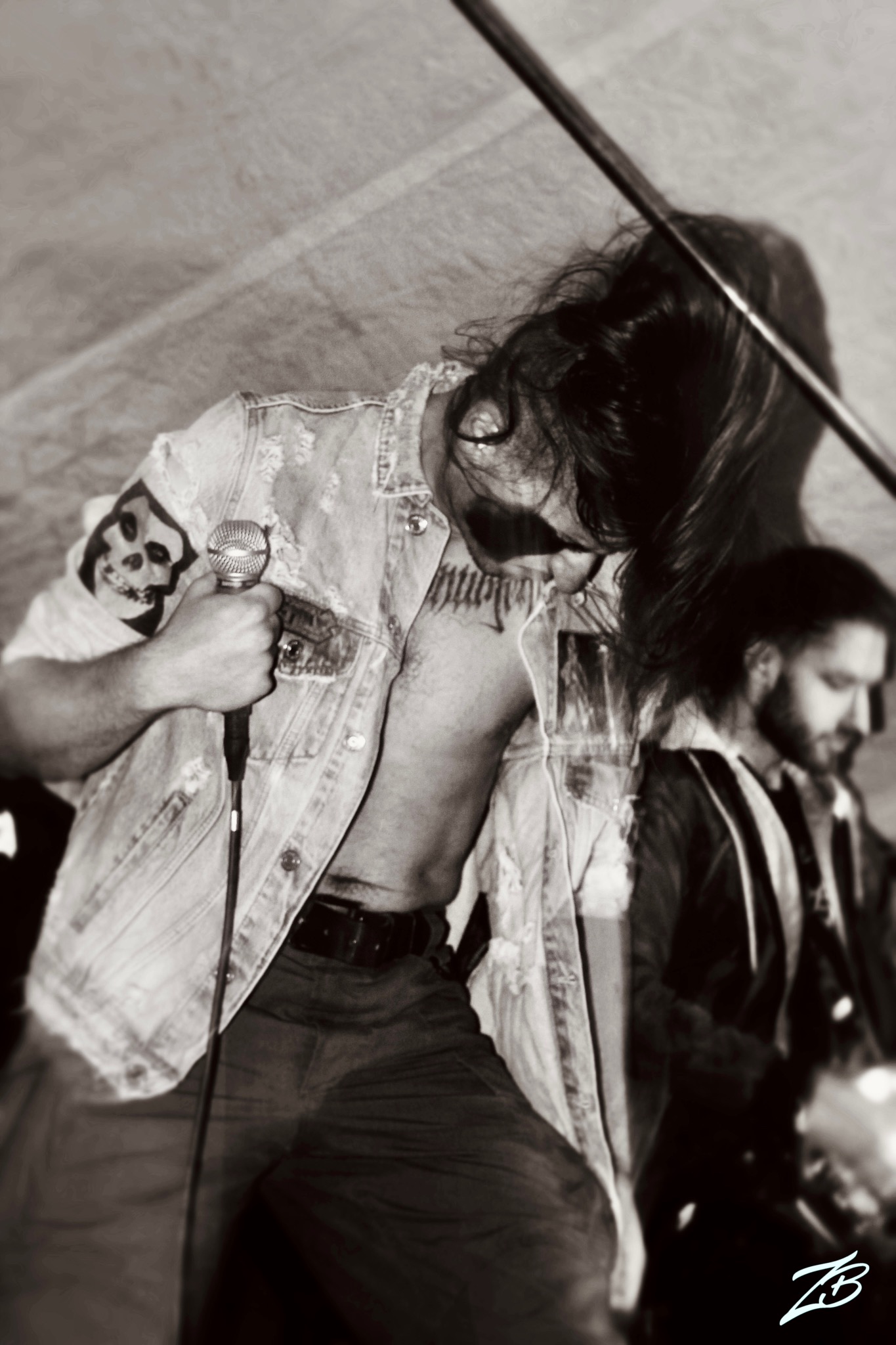 Czarno-białe zdjęcie wokalisty z długimi włosami, w dżinsowej kamizelce z naszywką czaszki, śpiewającego do mikrofonu podczas koncertu. W tle drugi muzyk.