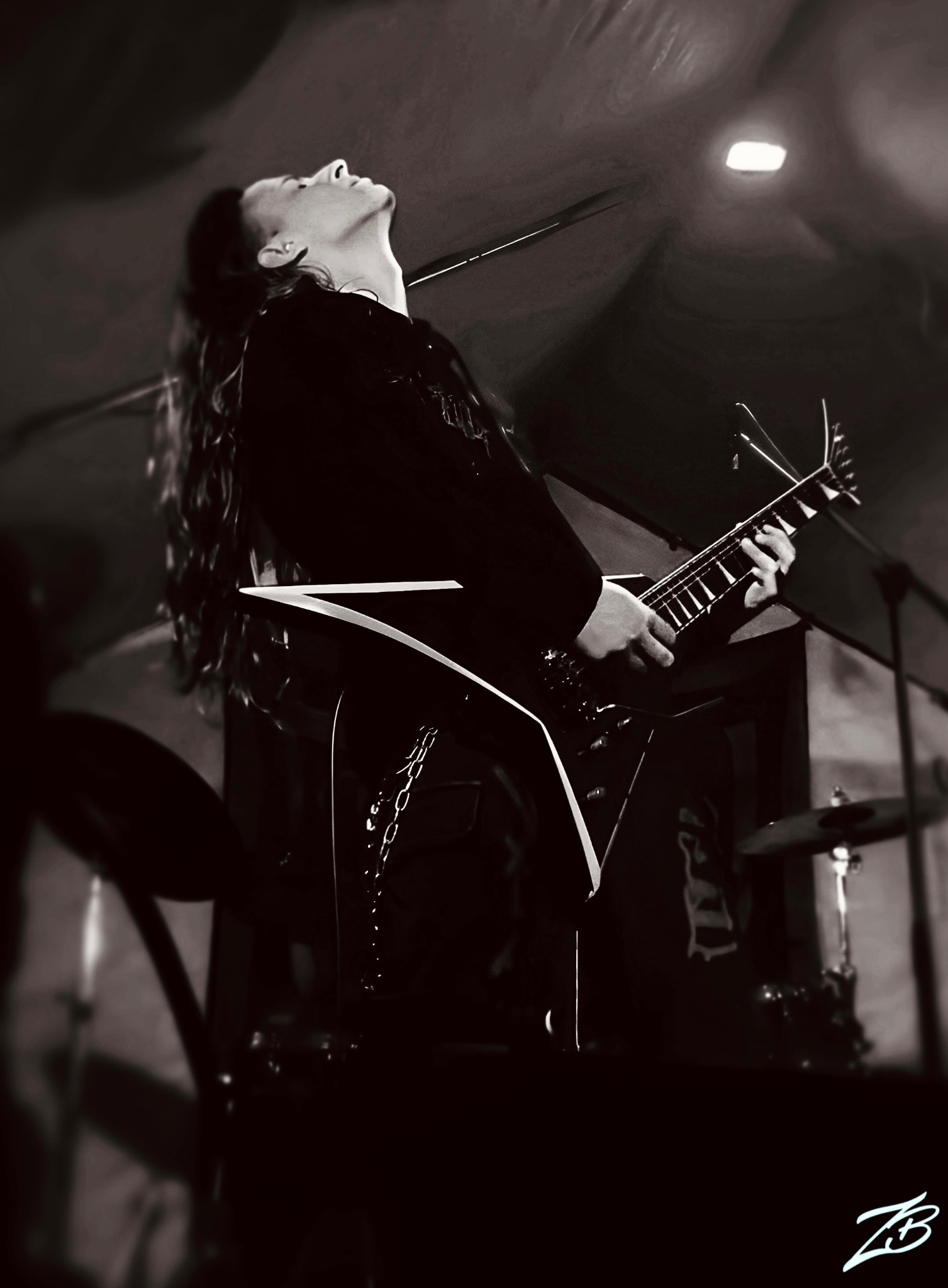 Czarno-białe zdjęcie gitarzysty z długimi włosami, grającego na gitarze typu V podczas występu na scenie. Widoczny fragment perkusji w tle.