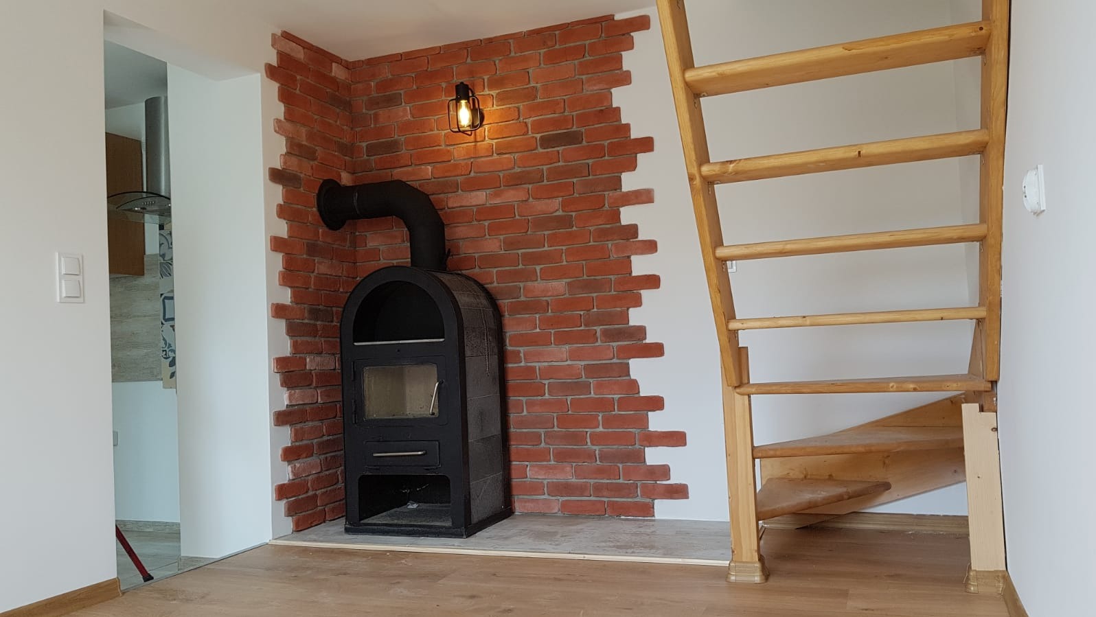 Ceglana ściana z kominkiem i oświetleniem w stylu loft, drewniane schody w jasnym wnętrzu. Dekoracyjne wykończenie ściany z cegły.