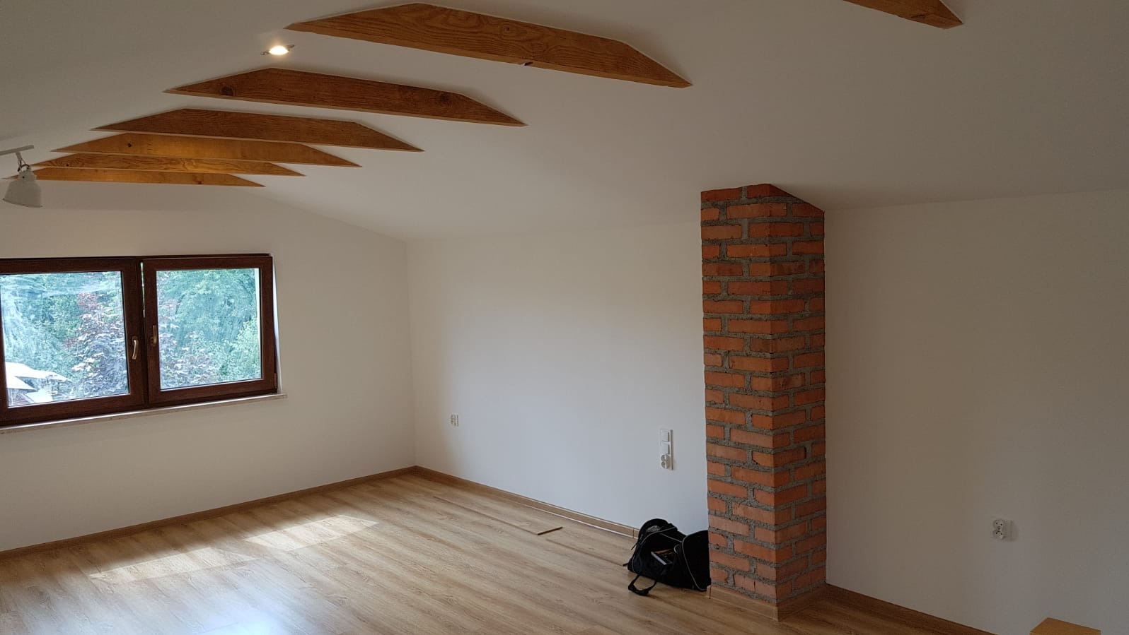 Poddasze po remoncie w Lubomi: drewniane belki stropowe, ściany pomalowane na biało, okno z widokiem na zieleń, komin z cegły, panele podłogowe.