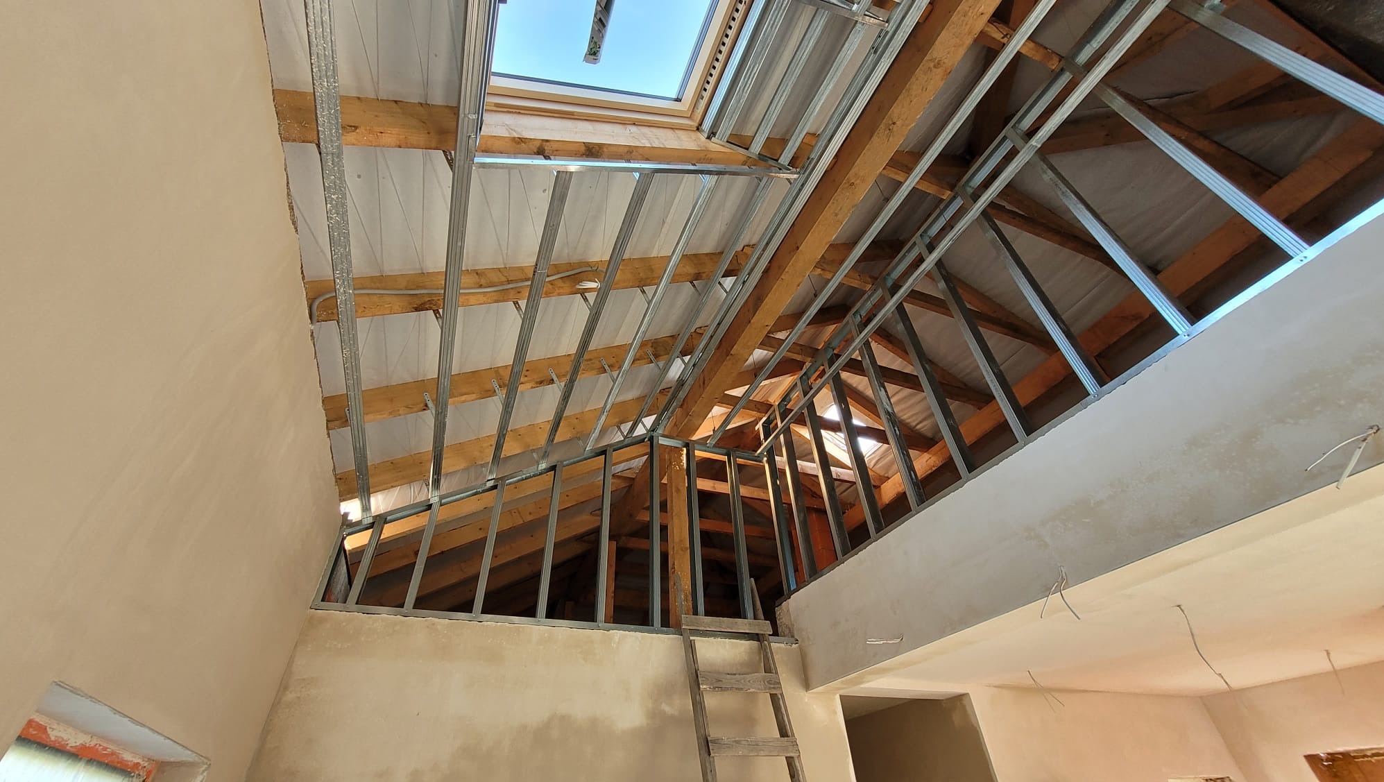 Poddasze w trakcie adaptacji: drewniana konstrukcja dachu z metalowym stelażem pod płyty g-k, fragment otynkowanej ściany i drabina. Widoczne okno dachowe.