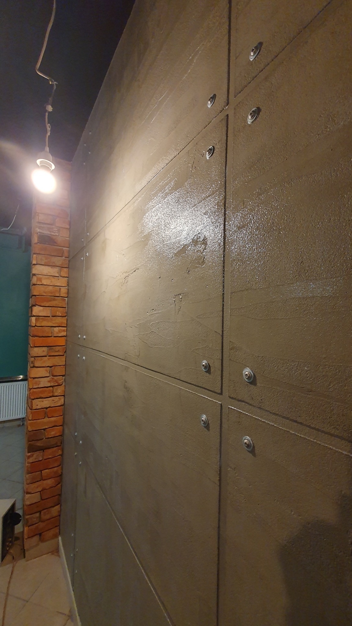 Ściana wykończona panelami imitującymi beton, połączonymi śrubami, z fragmentem ściany z cegły w tle. Widoczne oświetlenie sufitowe.