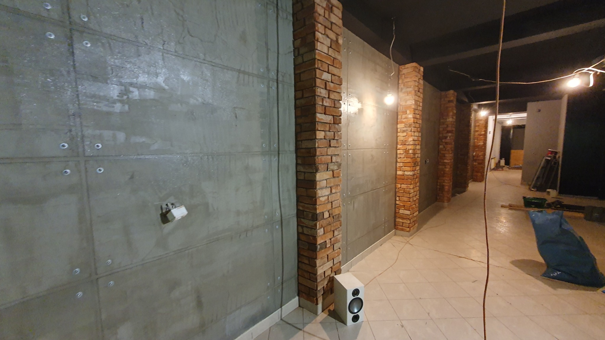 Surowa ściana z betonu architektonicznego z widocznymi śrubami, kontrastująca z ceglanymi filarami w industrialnym wnętrzu. Widoczny głośnik na podłodze.