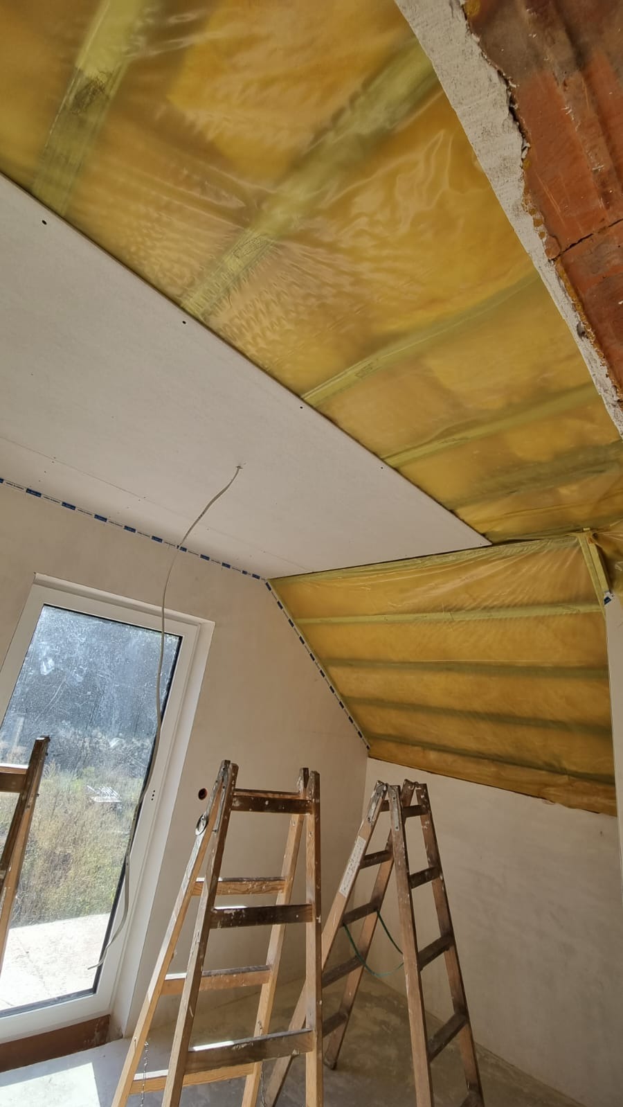 Poddasze w trakcie ocieplania: folia paroizolacyjna, częściowo zamontowana płyta gips-karton, okno i drabiny malarskie. Prace wykończeniowe w domu.