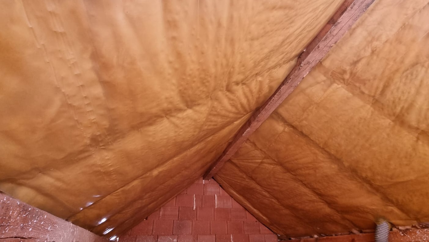 Poddasze ocieplone pianą PUR, widoczne krokwie i fragment muru z czerwonej cegły. Izolacja natryskowa o jednolitej strukturze pokrywa skośne powierzchnie dachu.