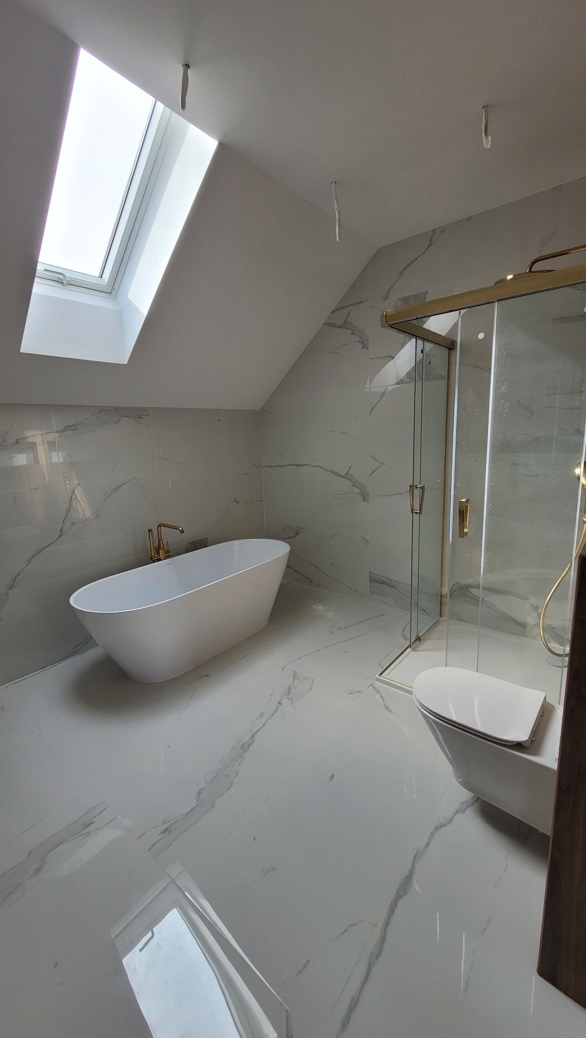 Jasna łazienka na poddaszu z marmurowymi płytkami, białą wanną z złotą armaturą i kabiną prysznicową ze złotymi ramami. Okno dachowe wpuszcza naturalne światło.