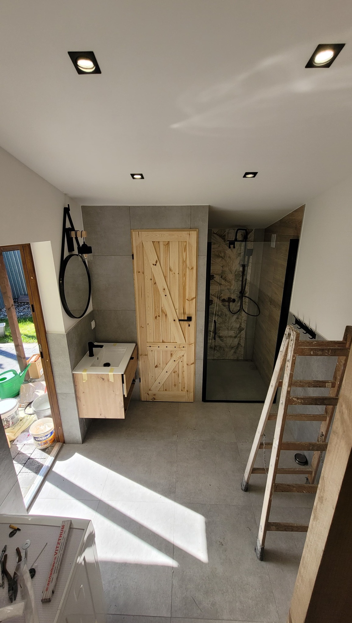 Wykończona łazienka z drewnianymi drzwiami i kabiną prysznicową w stylu rustykalnym. Widoczna umywalka z szafką i drabina malarska.