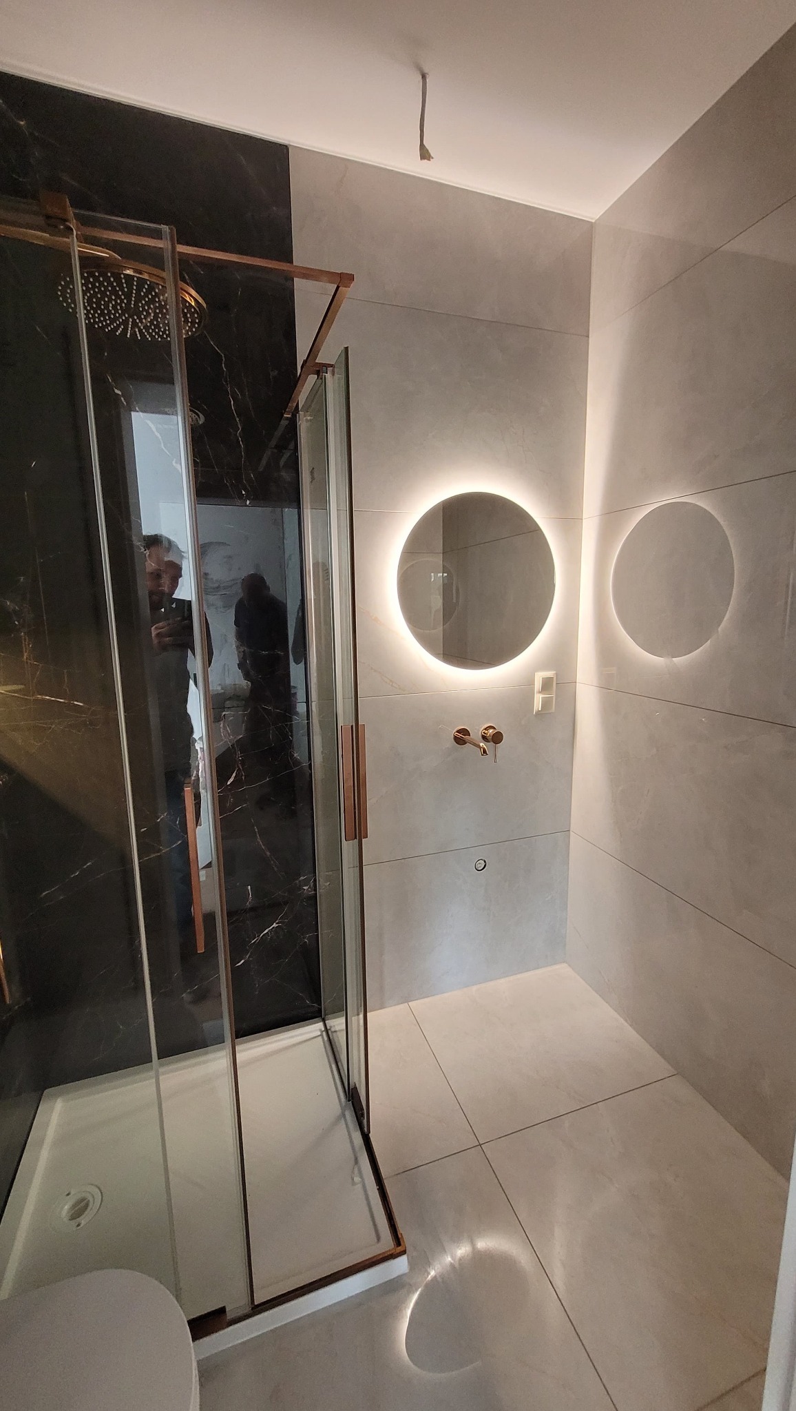 Nowoczesna łazienka z kabiną prysznicową typu walk-in, czarnymi płytkami imitującymi marmur i okrągłym lustrem z podświetleniem LED. Minimalistyczny design.