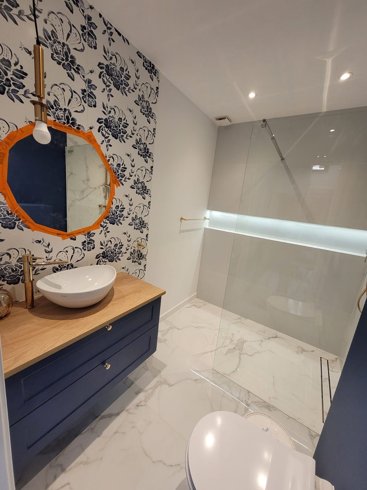 Nowoczesna łazienka z umywalką nablatową, szafką w kolorze granatowym i kabiną prysznicową typu walk-in. Na ścianie tapeta w kwiaty i okrągłe lustro z pomarańczową ramą.