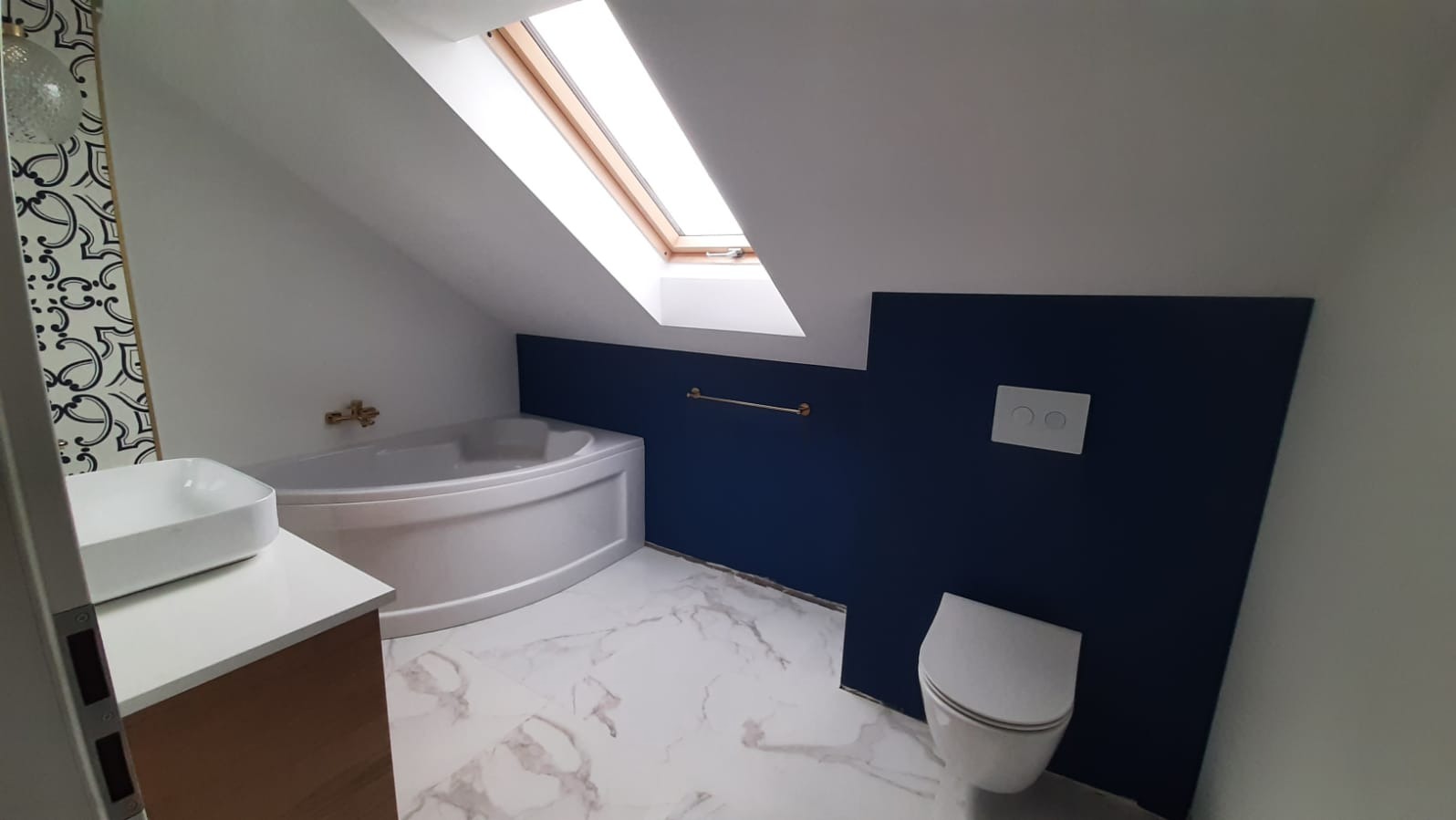 Nowoczesna łazienka na poddaszu z oknem dachowym, białą wanną narożną, umywalką nablatową i toaletą. Granatowa ściana z białym przyciskiem spłukującym. Marmurowe płytki podłogowe.