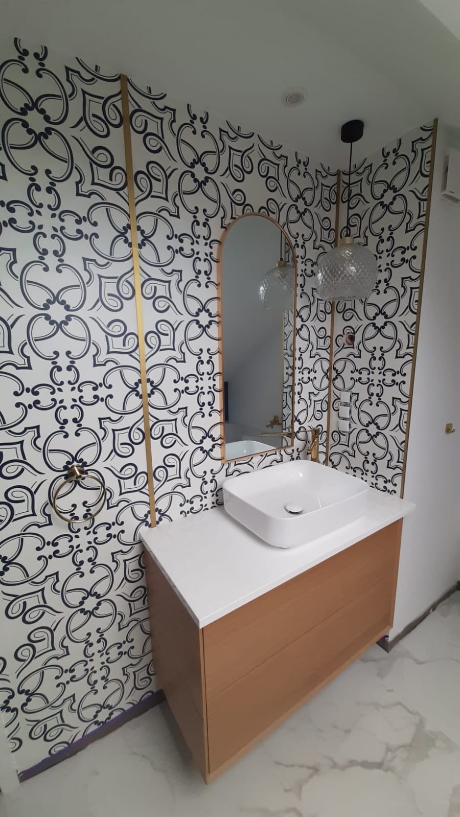 Wykończona łazienka z wzorzystą tapetą, złotymi listwami i nowoczesną umywalką nablatową. Lustro w złotej ramie i marmurowe płytki podłogowe.