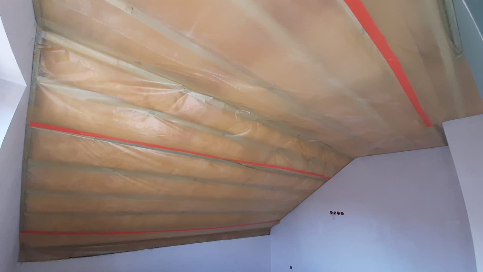 Ocieplone poddasze z widoczną izolacją termiczną i folią paroizolacyjną, zabezpieczone czerwoną taśmą. Widok z dołu na skośny sufit.