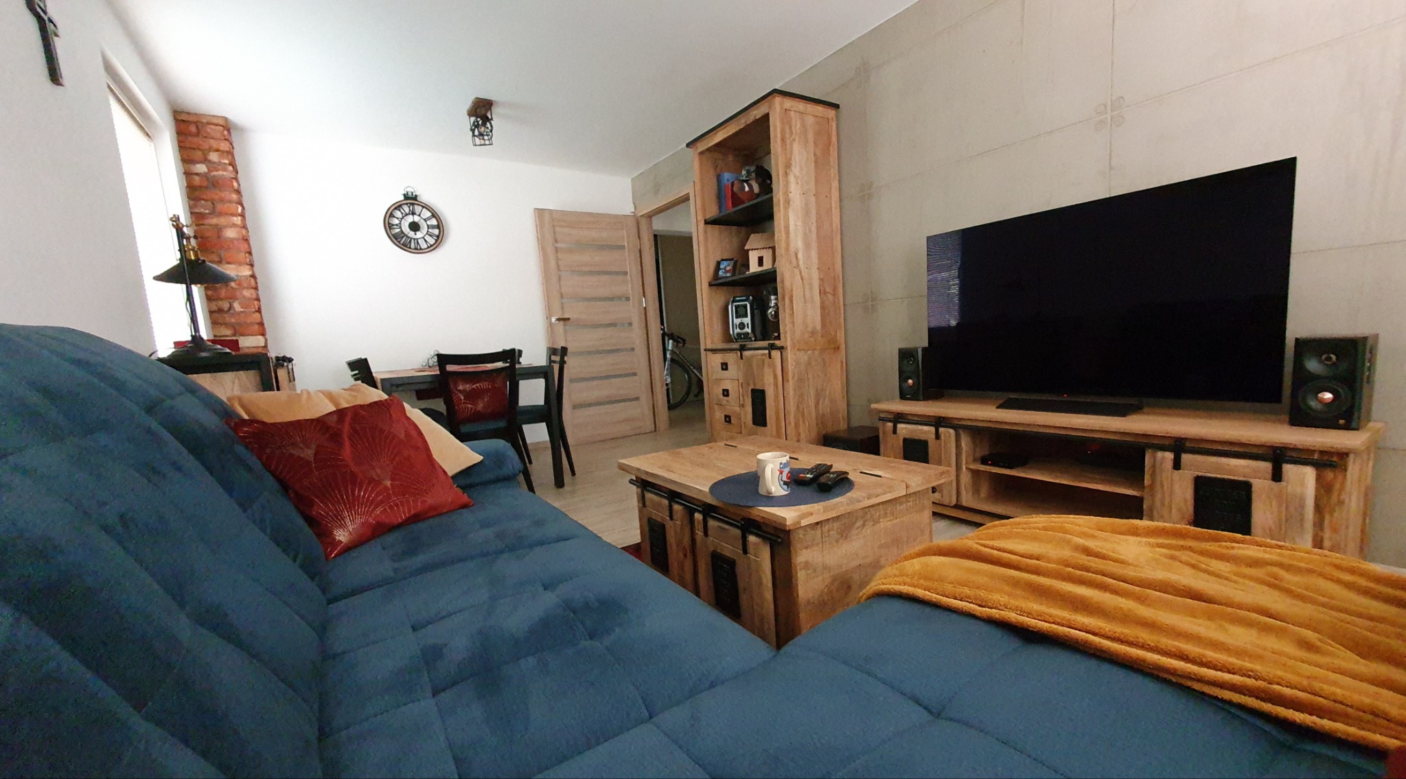 Jasne wnętrze salonu z drewnianymi meblami: szafka RTV, regał, stolik kawowy. Niebieska sofa z poduszkami i kocem. Telewizor na ścianie. Widok na jadalnię.