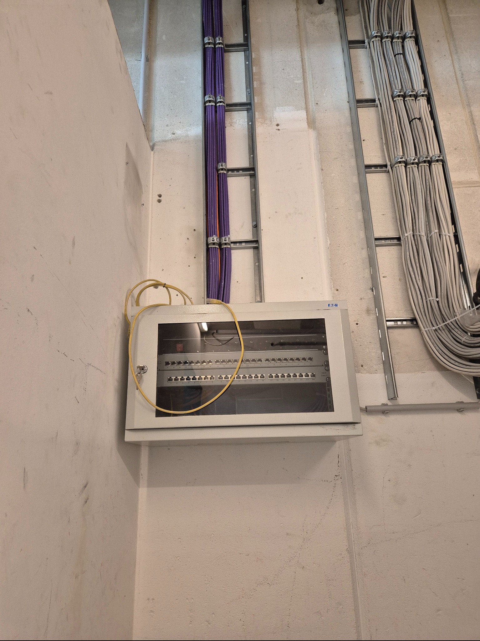 Szara szafka elektryczna marki Eaton zamontowana na ścianie, z widocznymi kablami w korytkach. Kable fioletowe i szare. Widoczny zamek w szafce.