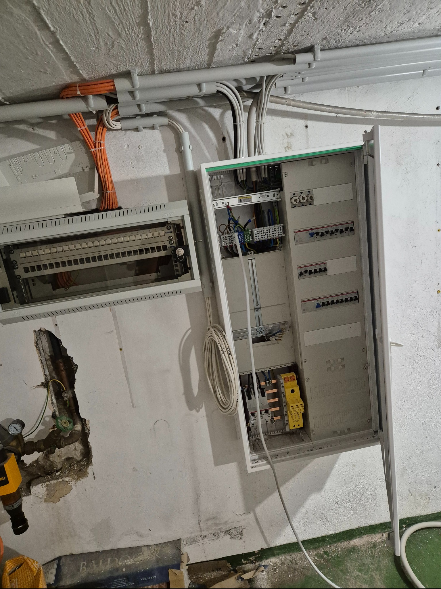 Otwarta skrzynka elektryczna z widocznymi przewodami i bezpiecznikami na tle ściany z instalacjami. Widoczne kable w peszlach. Prace elektryczne w surowym wnętrzu.