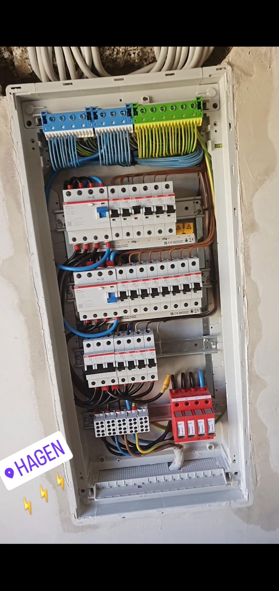 Rozdzielnica elektryczna z bezpiecznikami i przewodami w kolorach: niebieskim, zielonym, brązowym i czarnym, zamontowana w ścianie. Widoczne zabezpieczenia przeciwprzepięciowe.