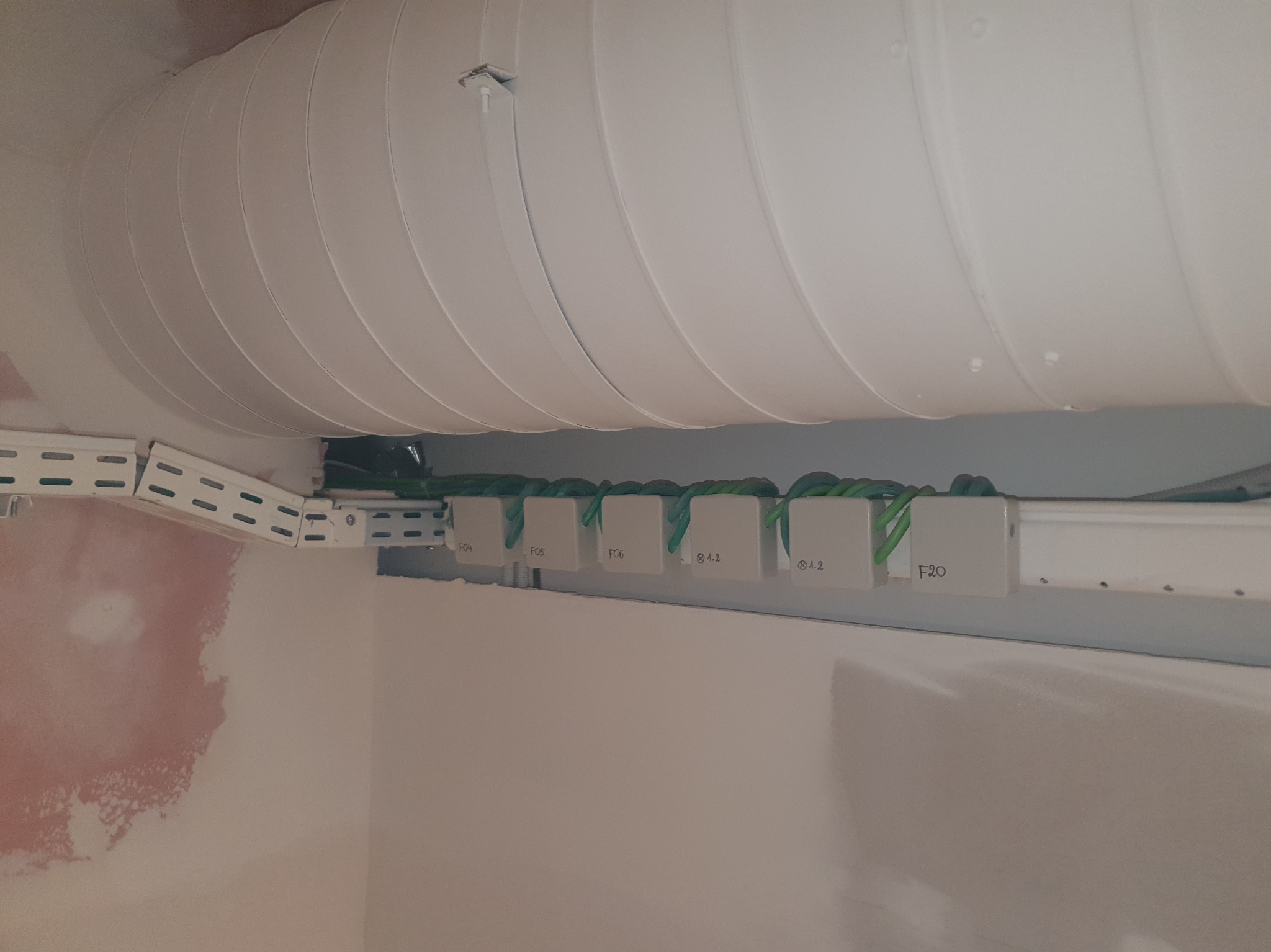 Białe puszki z oznaczeniami F04, F05, F06, 8A.2 i F20, zamontowane na metalowej szynie pod sufitem, z widoczną instalacją wentylacyjną. Ściana w tle z śladami gładzi.