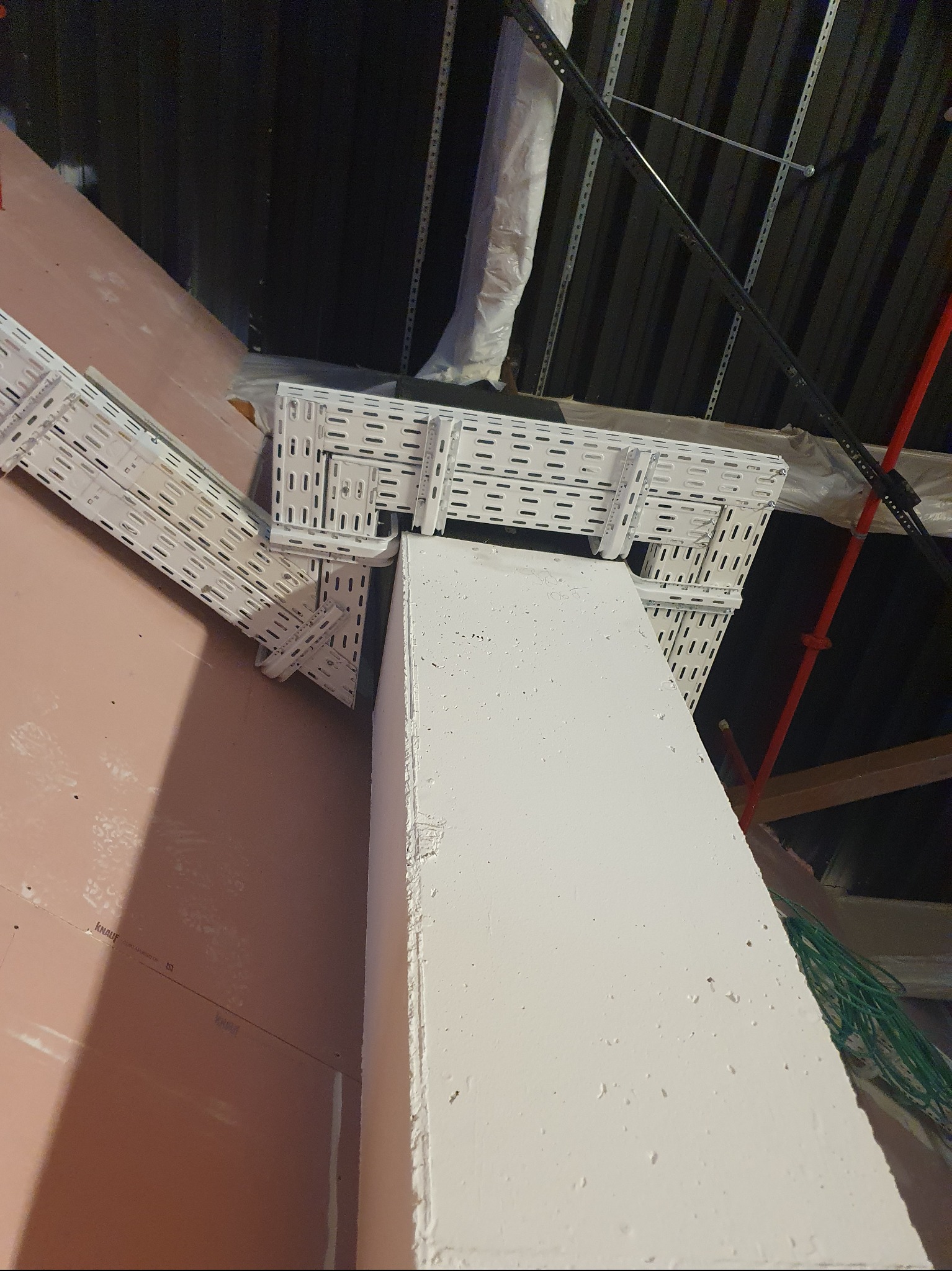 Białe korytka kablowe zamontowane na ścianie i słupie w surowym wnętrzu. Widoczne przewody i elementy konstrukcyjne. Ujęcie z dołu, podkreślające perspektywę.