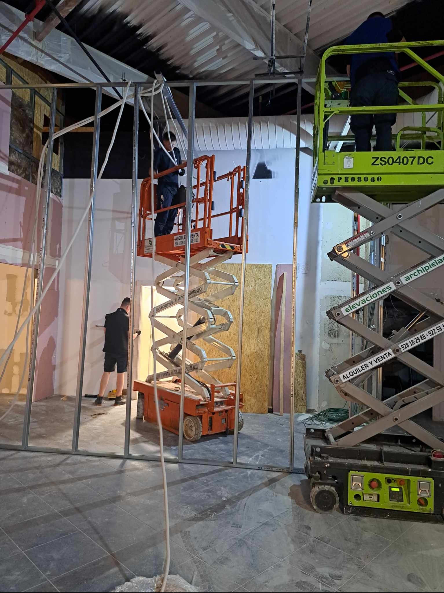 Prace wewnątrz budynku: elektryk na podnośniku układa przewody pod sufitem. Widoczne metalowe profile ścian działowych i drugi podnośnik. Etap prac wykończeniowych.