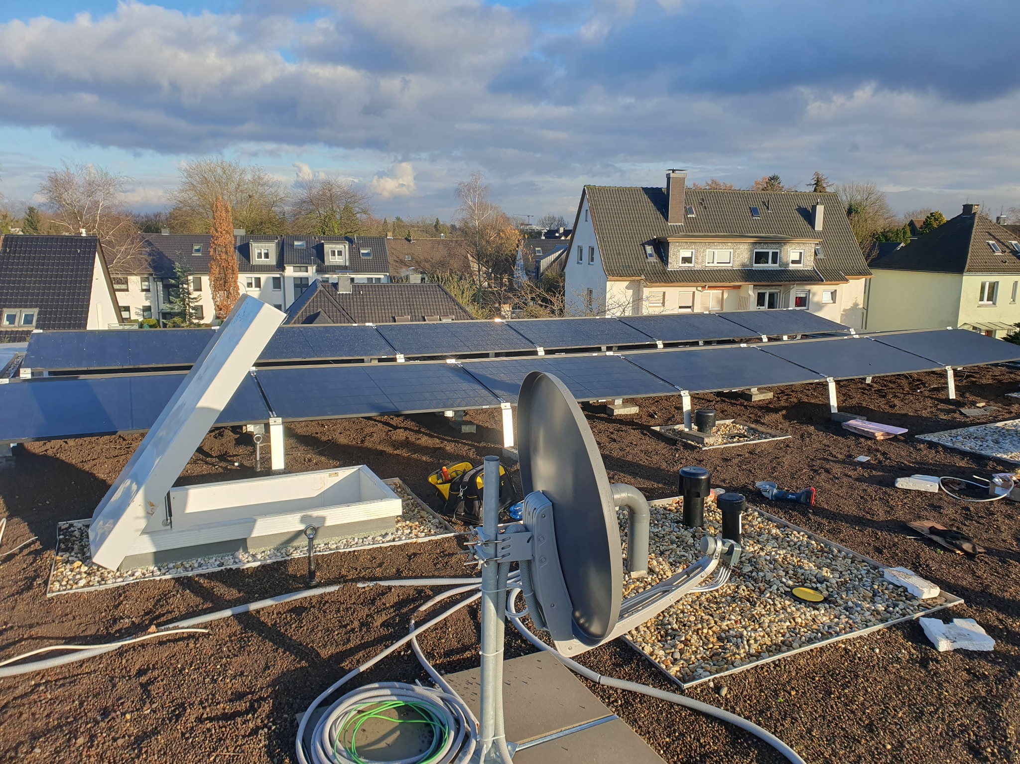 Instalacja paneli słonecznych na dachu z anteną satelitarną. Widoczne narzędzia i kable. Tło: zabudowa jednorodzinna i błękitne niebo z chmurami.