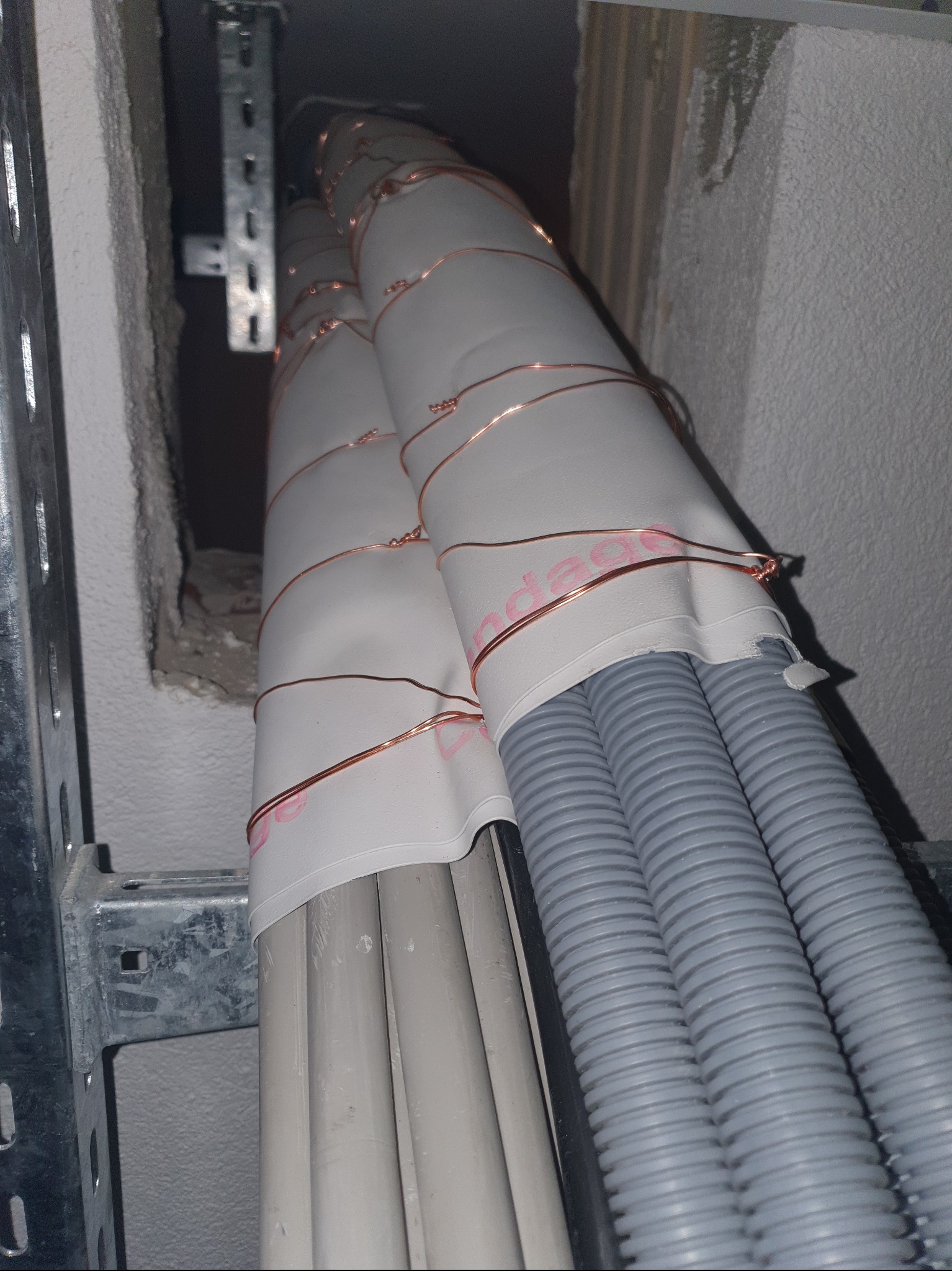 Szare rury karbowane i gładkie, zabezpieczone białą taśmą i miedzianym drutem, prowadzone wzdłuż metalowej konstrukcji w przestrzeni między ścianami.