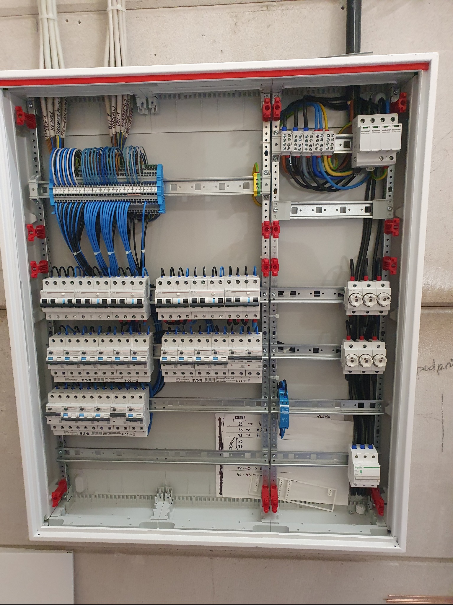 Rozdzielnica elektryczna w białej obudowie, z licznymi bezpiecznikami, przewodami w kolorach niebieskim i czarnym, zamontowana na ścianie. Widoczne oznaczenia na przewodach.