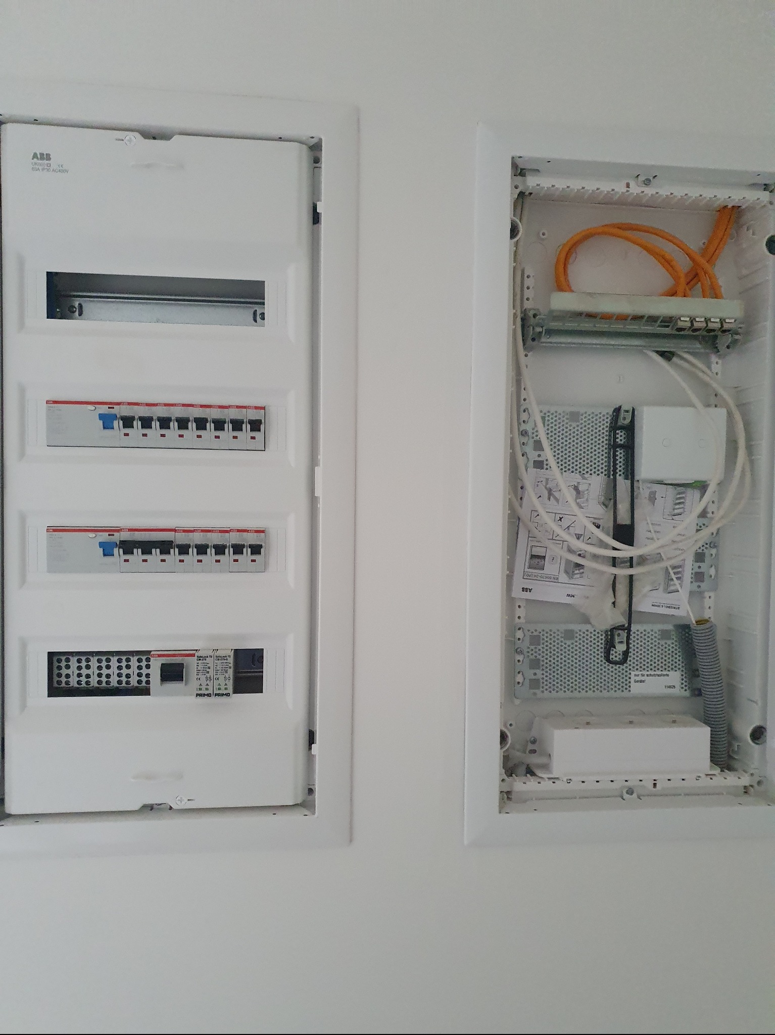 Dwie białe skrzynki elektryczne w ścianie: jedna z bezpiecznikami, druga z kablami i instrukcjami. Widok z bliska na instalację elektryczną wewnątrz budynku.