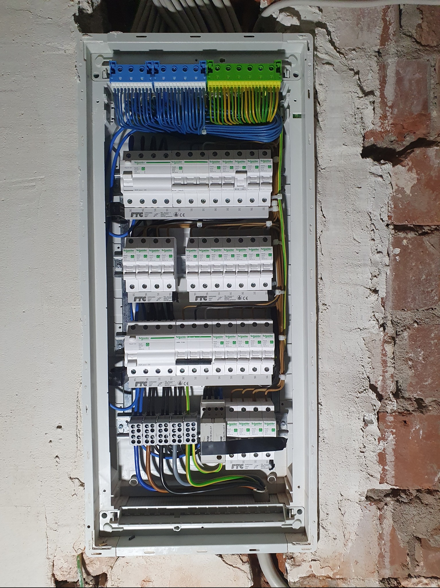 Rozdzielnica elektryczna Schneider z licznymi zabezpieczeniami, zamontowana na ścianie z surową cegłą i fragmentami tynku. Widoczne kable w różnych kolorach.